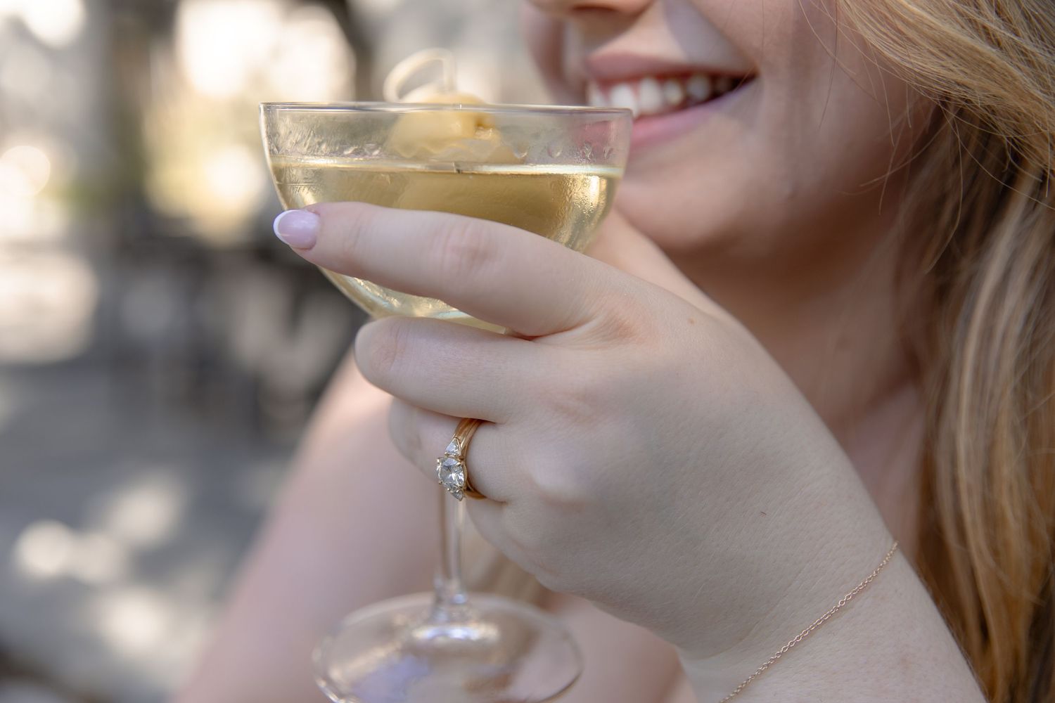 Someone holds a glass of white wine in an outdoor setting with dappled sunlight filtering through trees.