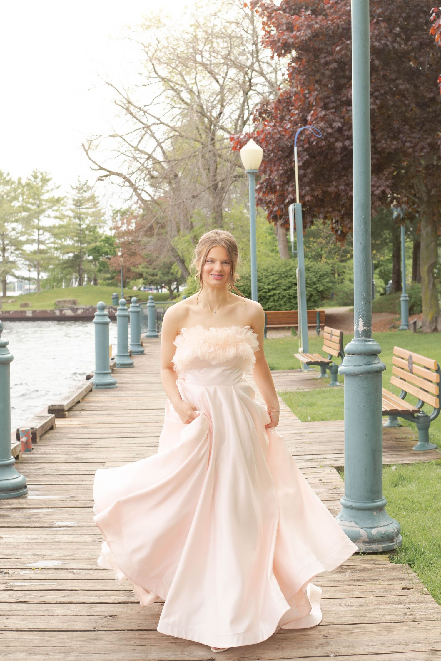 A flowing blush-colored gown twirls on a waterfront boardwalk with vintage-style lampposts.