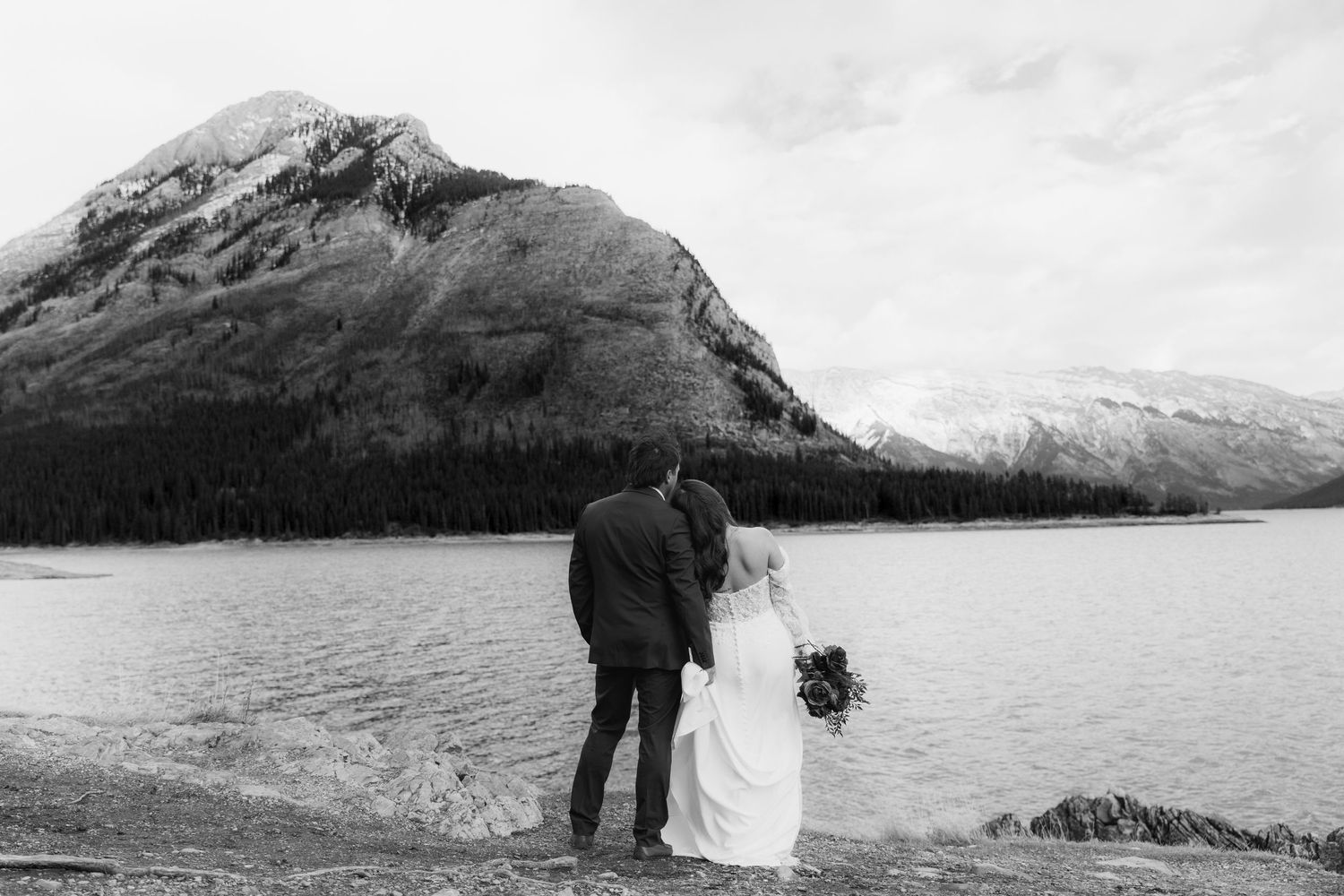 Couple in wedding attire embracing by a serene mountain lake with dramatic mountain landscape views.