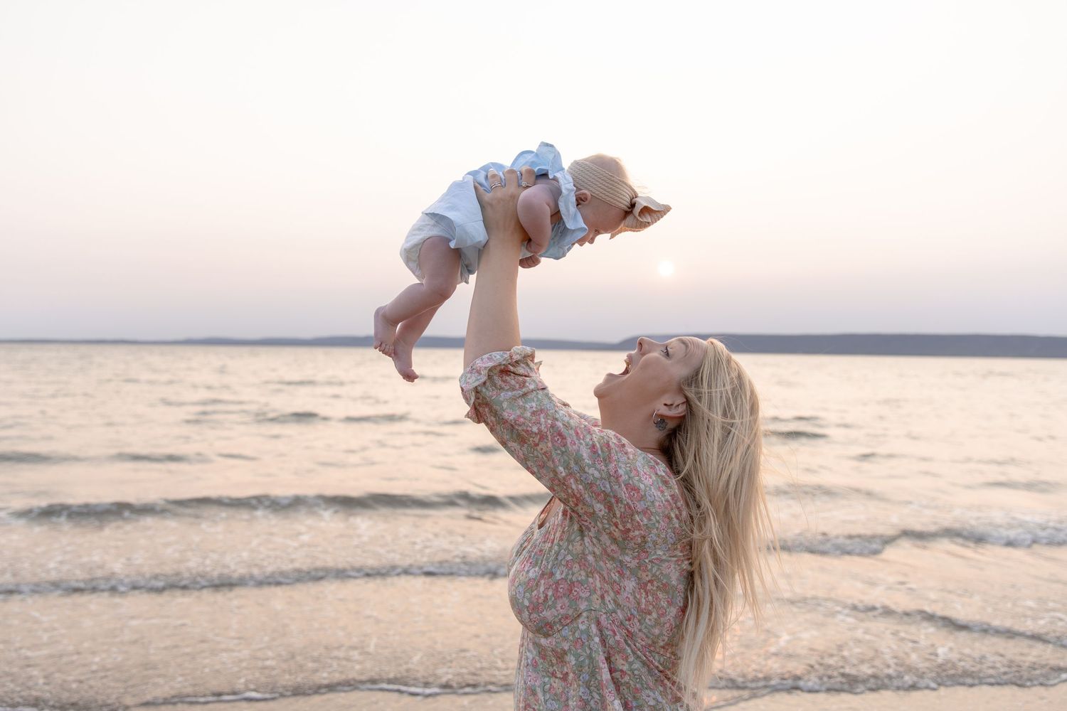 A playful moment at the beach as someone lifts a baby up against the sunset sky over the water.