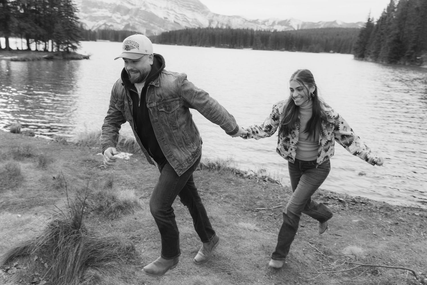 A couple runs playfully along a lakeside path with snow-capped mountains visible in the background.