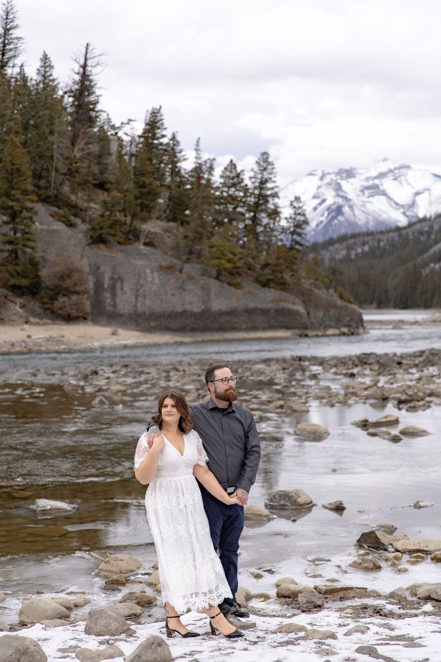 A couple stands together on a rocky riverbank with snow-capped mountains and evergreen forest in the background.