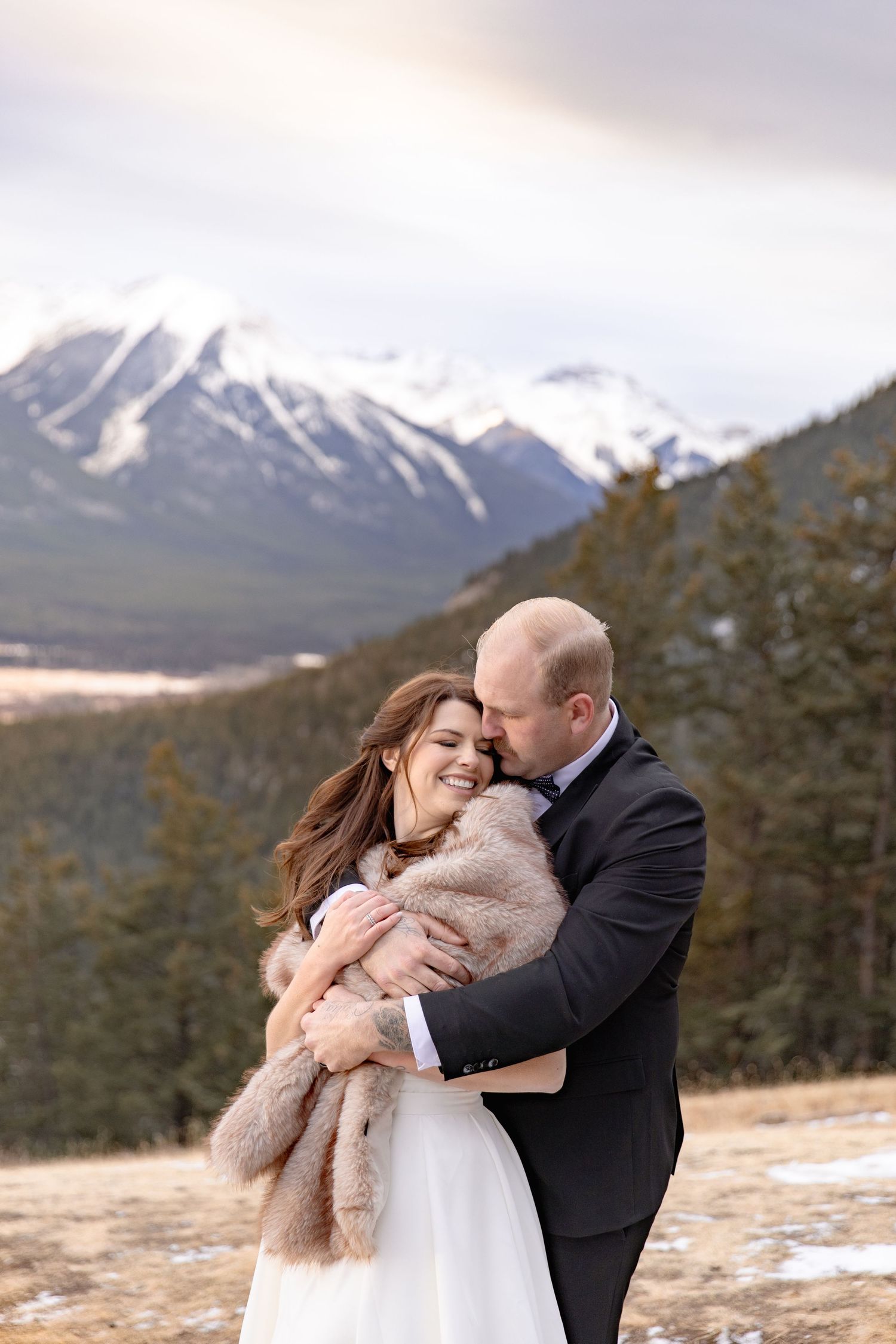 A romantic embrace between a couple in winter attire against a snowy mountain backdrop in the wilderness.