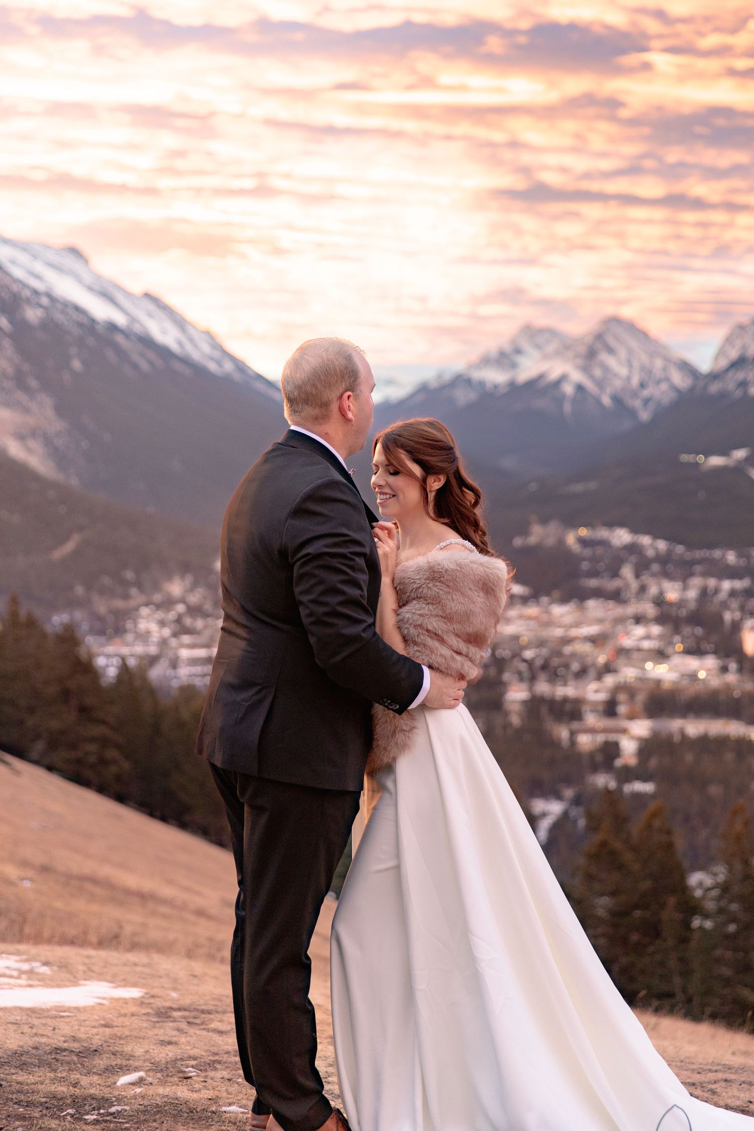 A couple in wedding attire shares an intimate moment against a backdrop of snow-capped mountains at sunset.