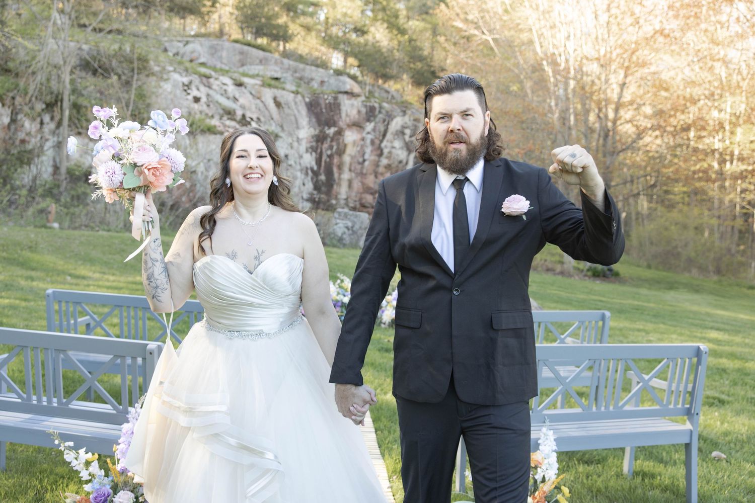 A couple celebrates joyfully at their outdoor wedding ceremony with blue wooden chairs and rocky backdrop at sunset.