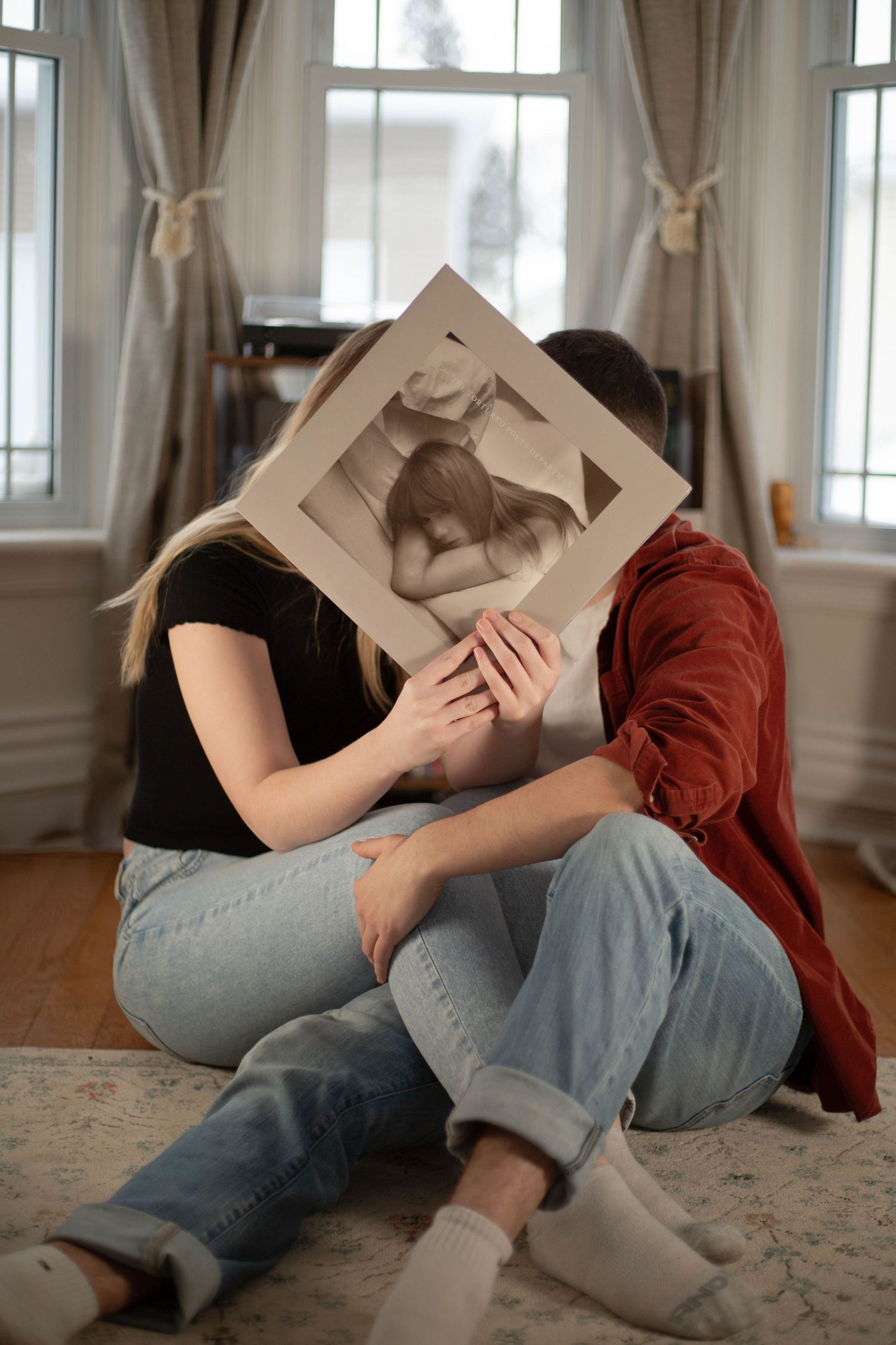 Indoor portrait with a vinyl record held up covering their faces, creating a playful composition near curtained windows.