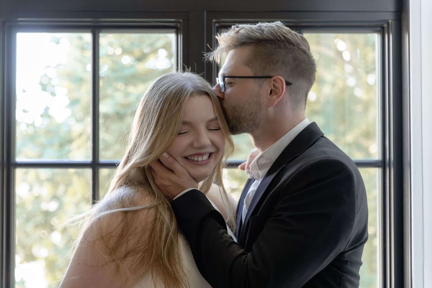 Happy couple in formal attire share an intimate moment near a bright window.