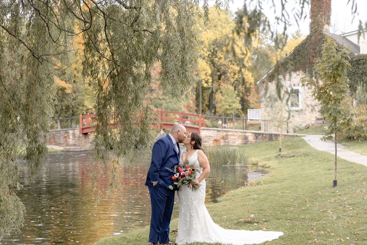 A couple shares a romantic moment under willow trees by a tranquil lake during their outdoor wedding photoshoot.