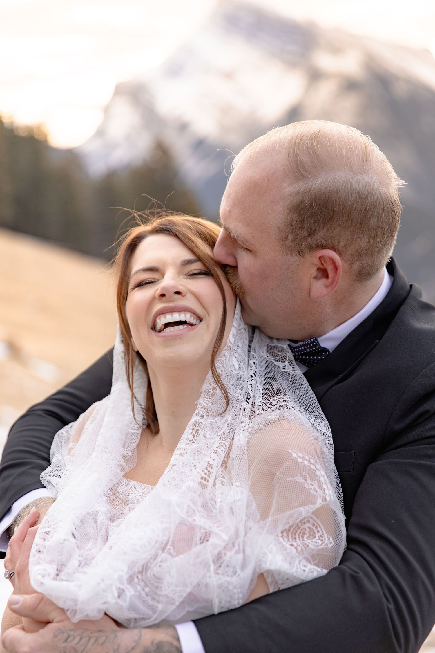A romantic wedding couple embraces and shares joyful laughter together in an outdoor setting during golden hour.