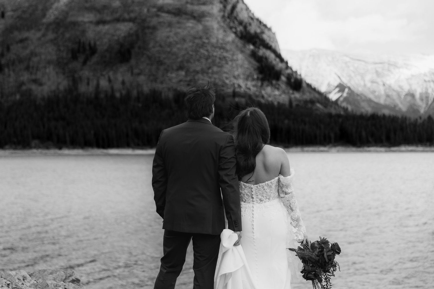 Wedding couple poses against dramatic mountain landscape with snow-capped peaks and lake in background.