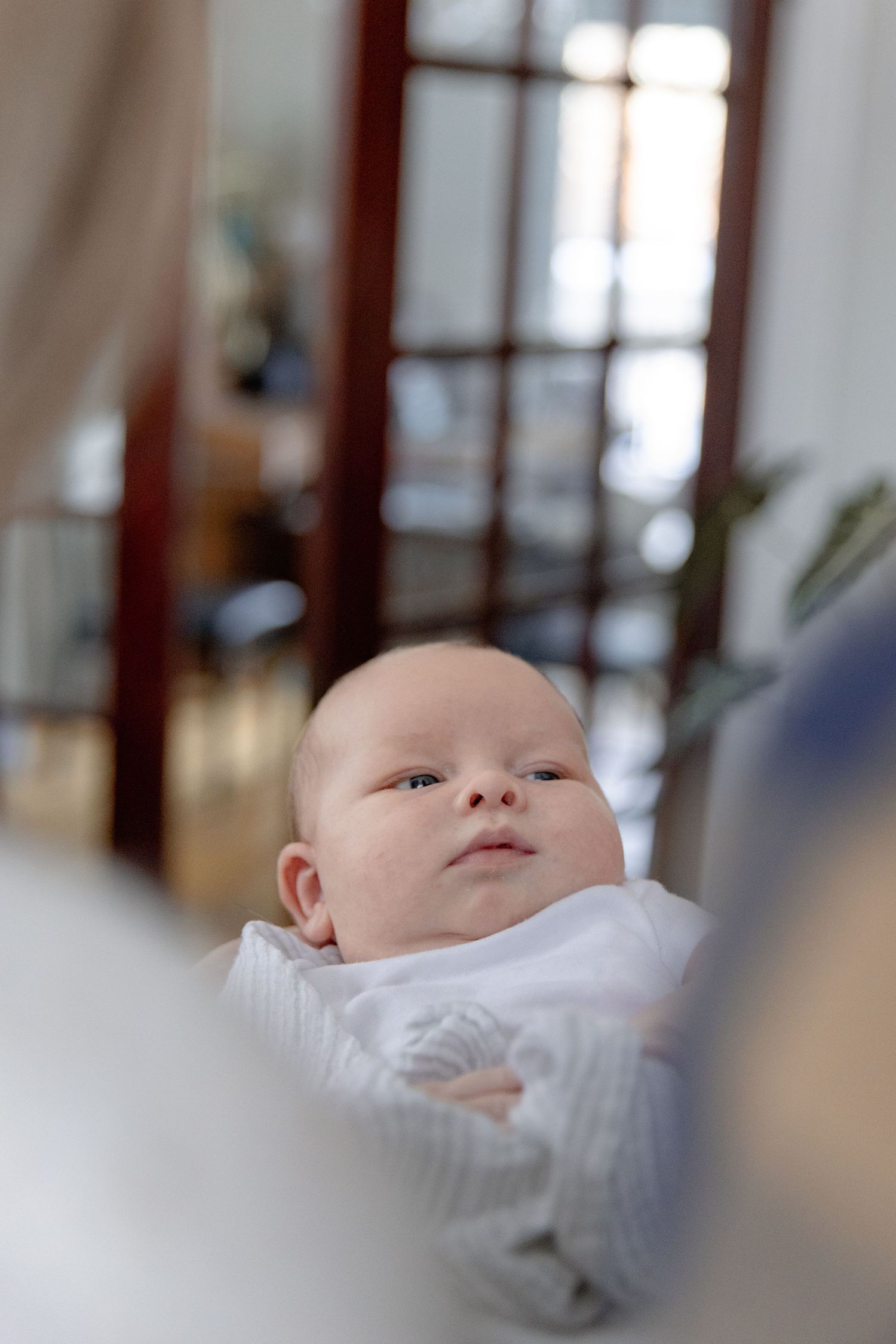 A baby in white clothing lies peacefully while looking upward in a soft-focus indoor setting.