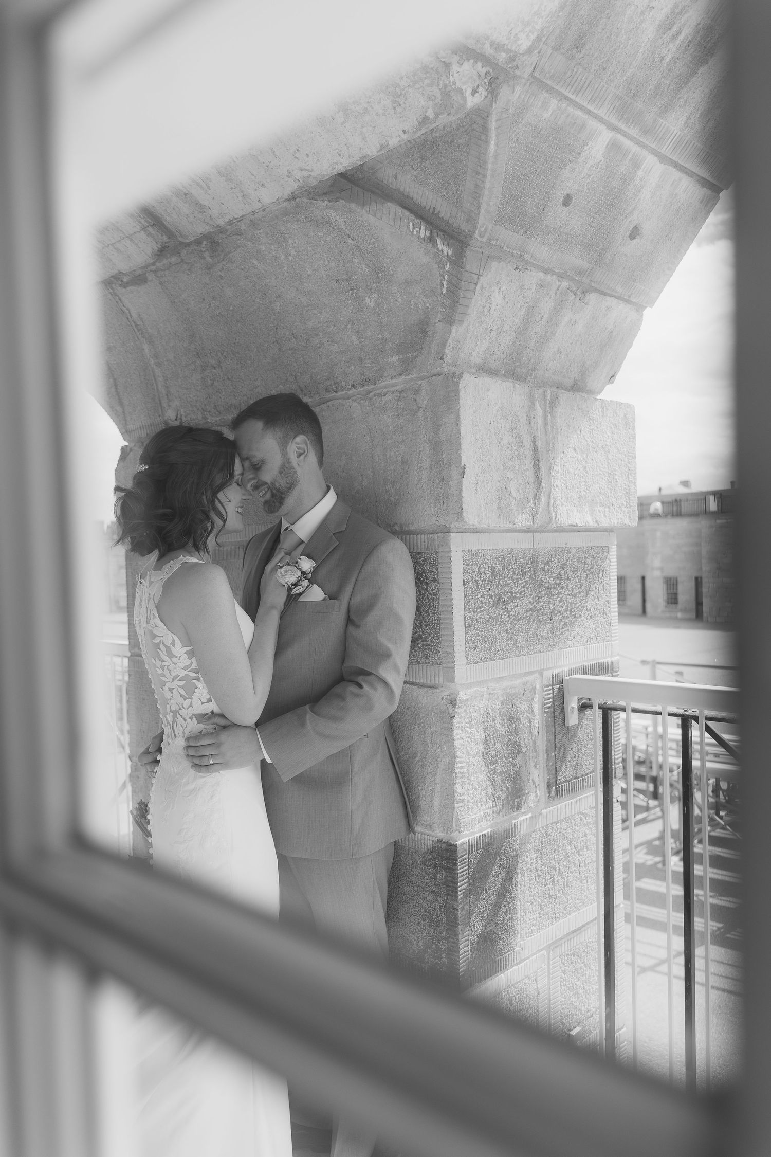Black and white wedding photo of bride and groom embracing on a stone balcony, captured through a window frame.