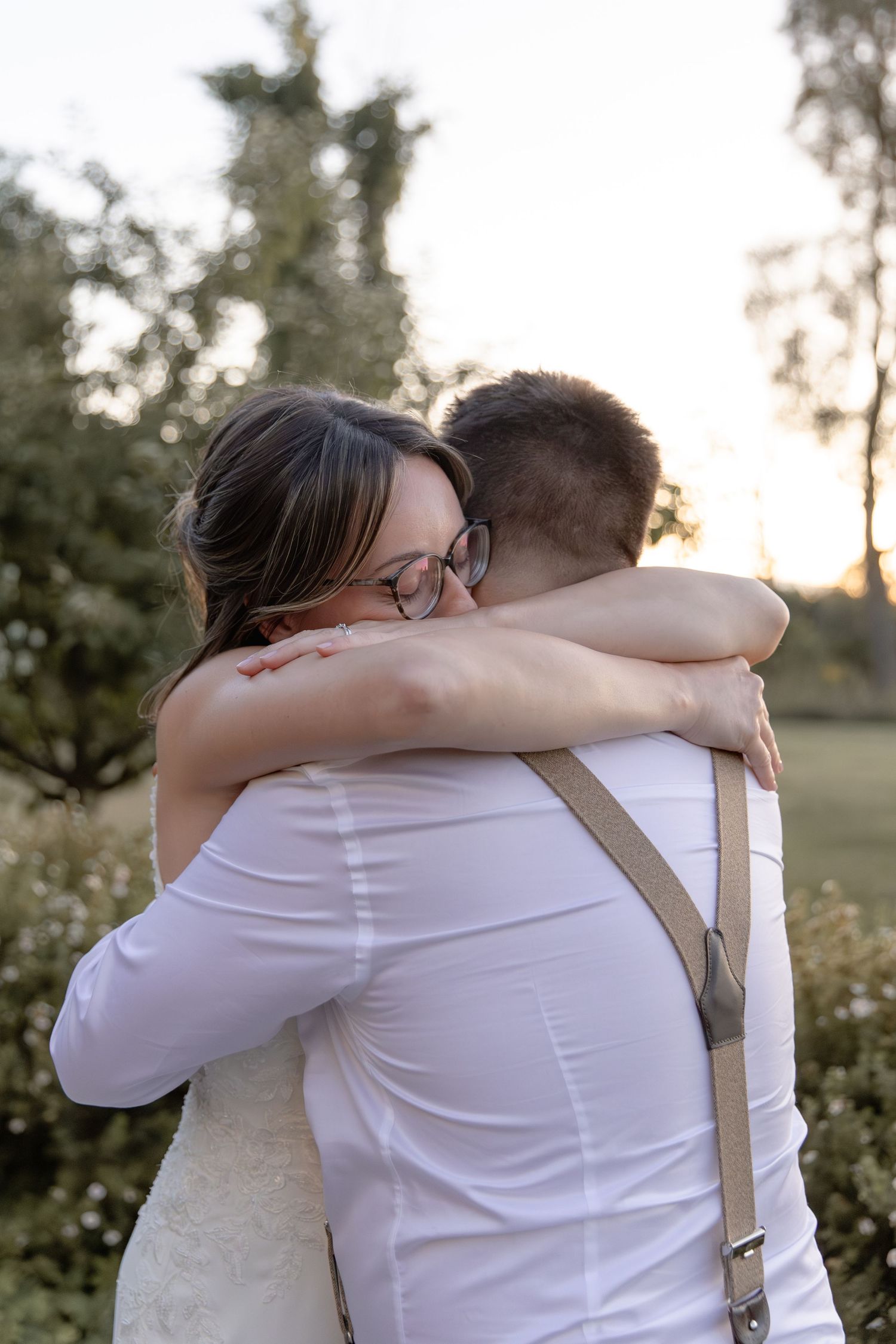 Two people wearing white embrace in an outdoor setting during sunset with trees in the background.