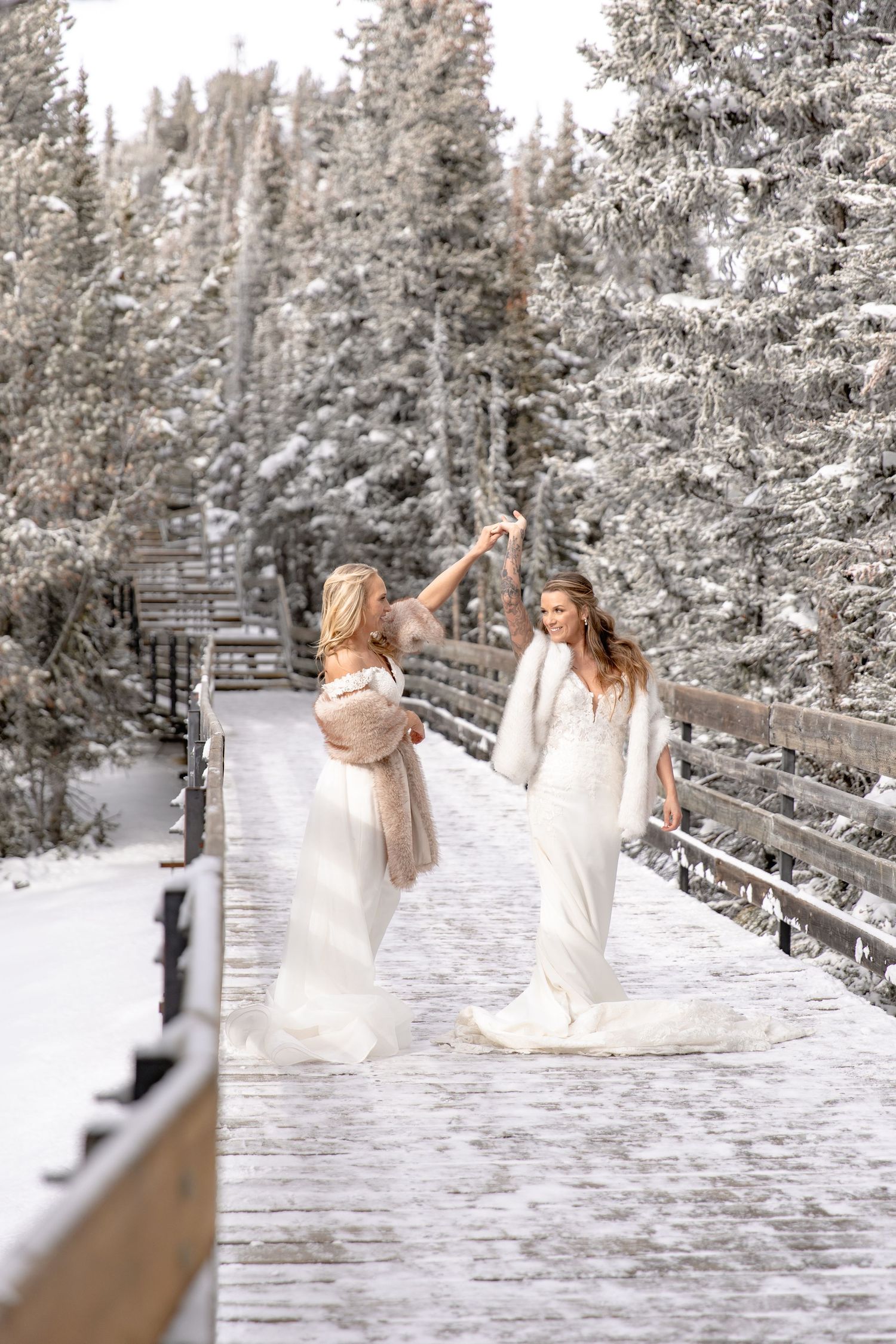 Two women in flowing white dresses twirl and dance playfully on a snow-covered boardwalk in a winter forest.