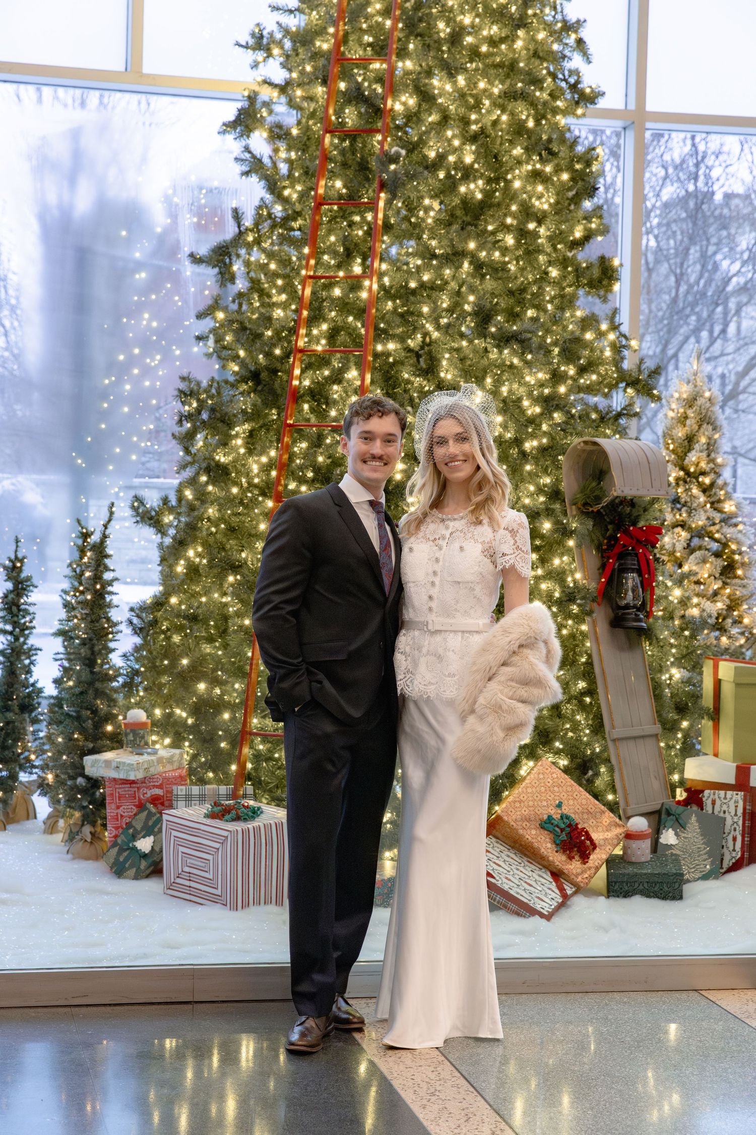 Couple poses in front of large Christmas tree with snowy winter scene visible through windows in elegant venue.