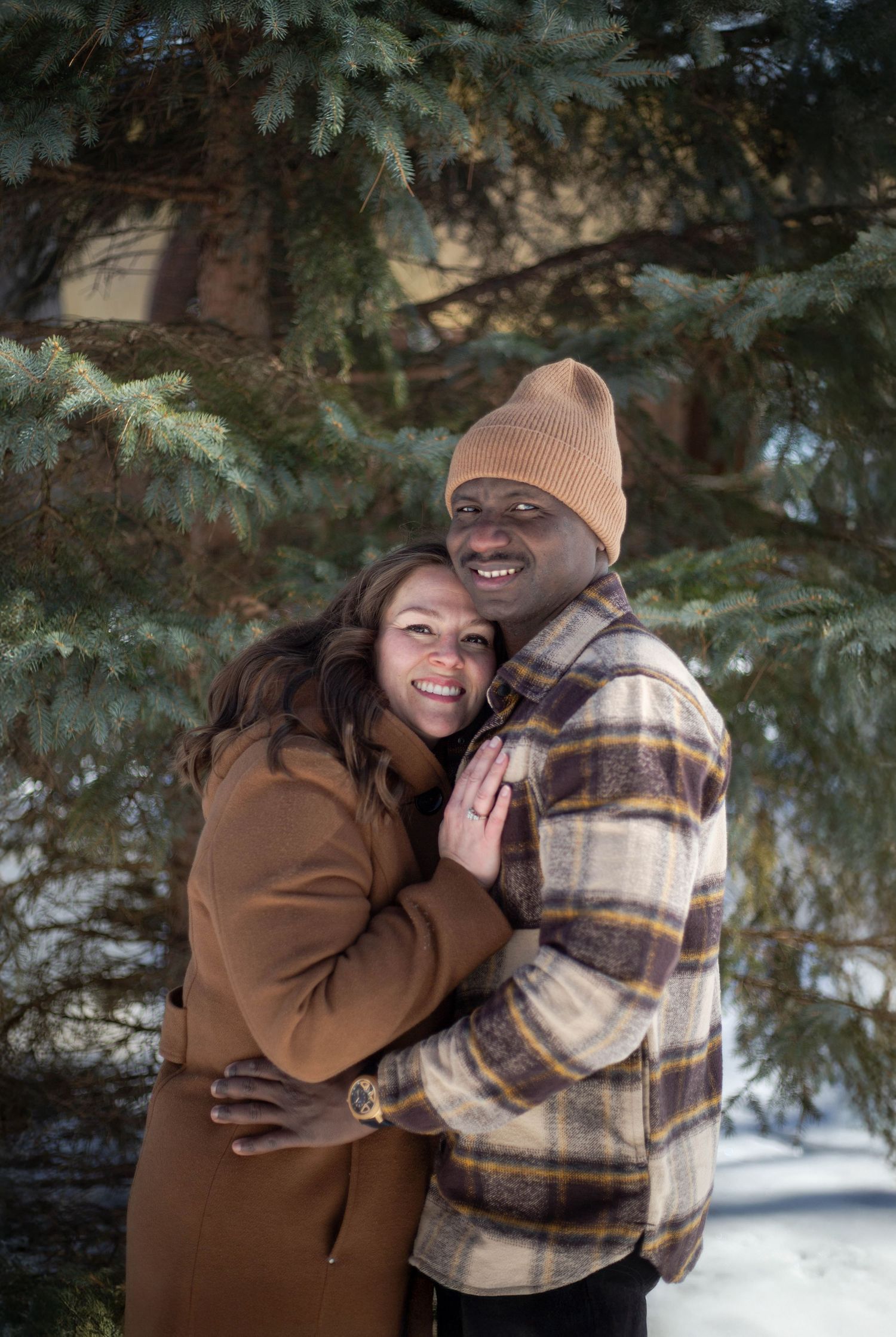 A couple shares a loving embrace while standing among snow-covered evergreen trees.
