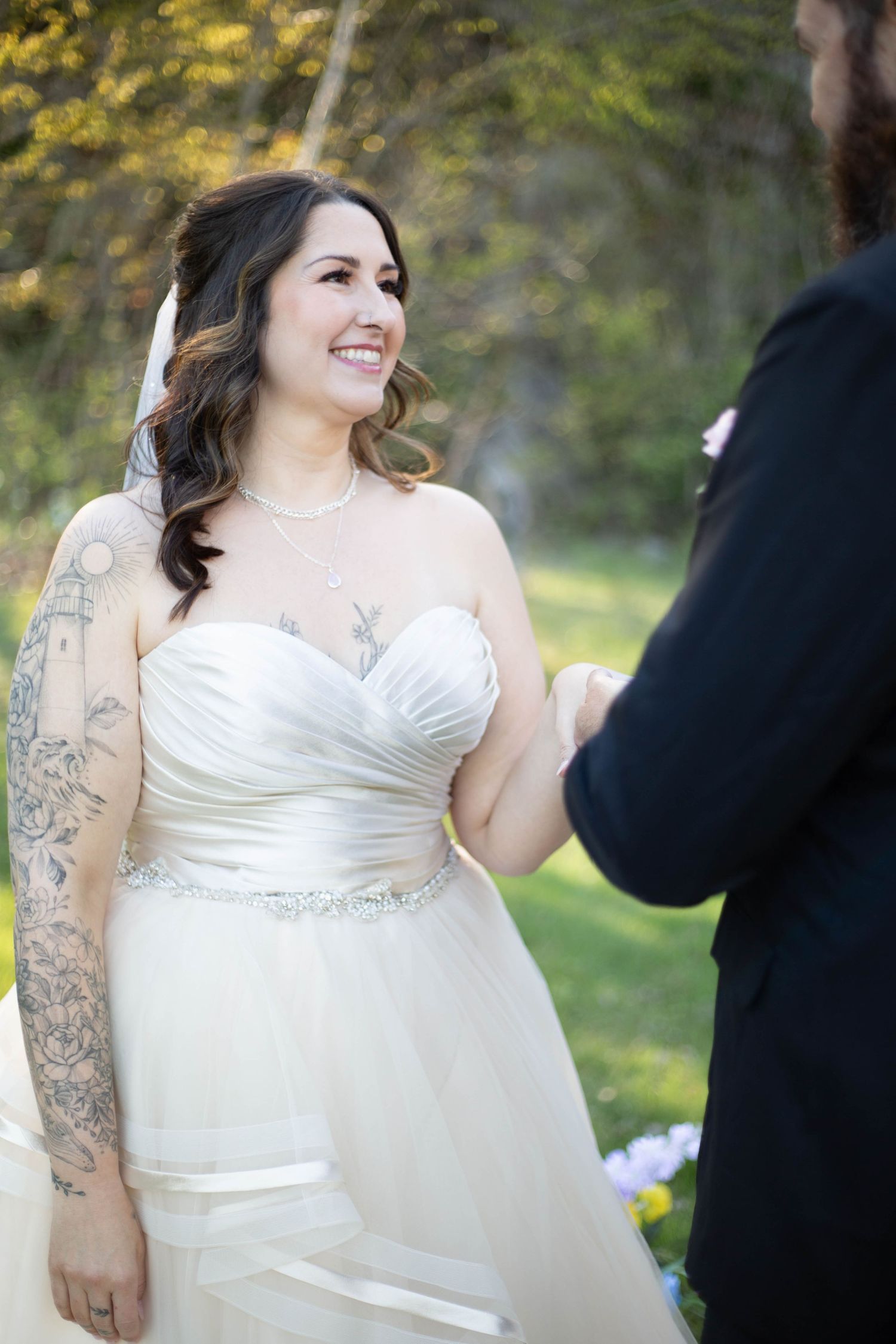 A bride wearing a strapless white wedding gown with beaded details and sleeve tattoos smiles during an outdoor ceremony.