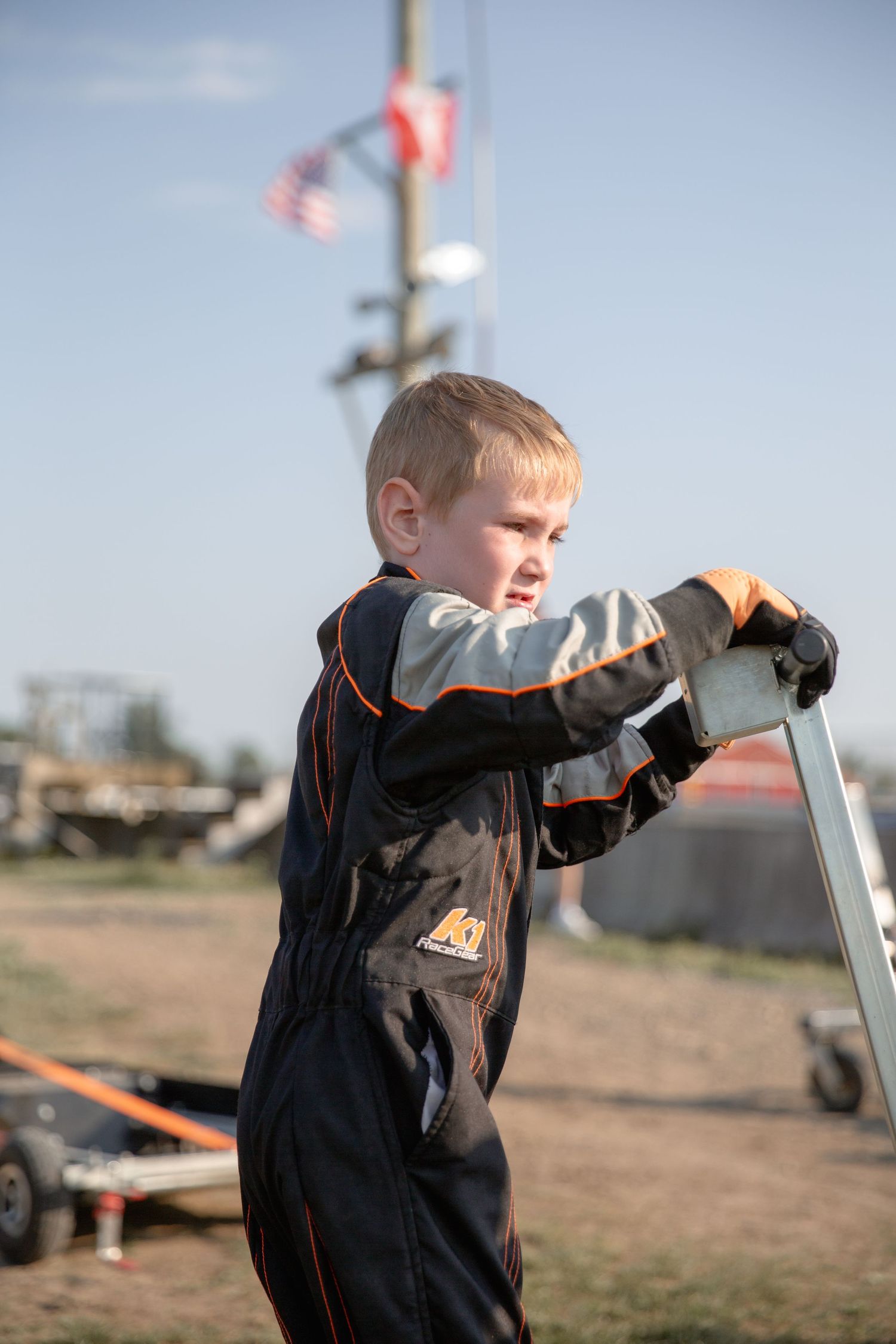 Young child in a navy coverall jumpsuit working on outdoor equipment with an American flag flying in the background.