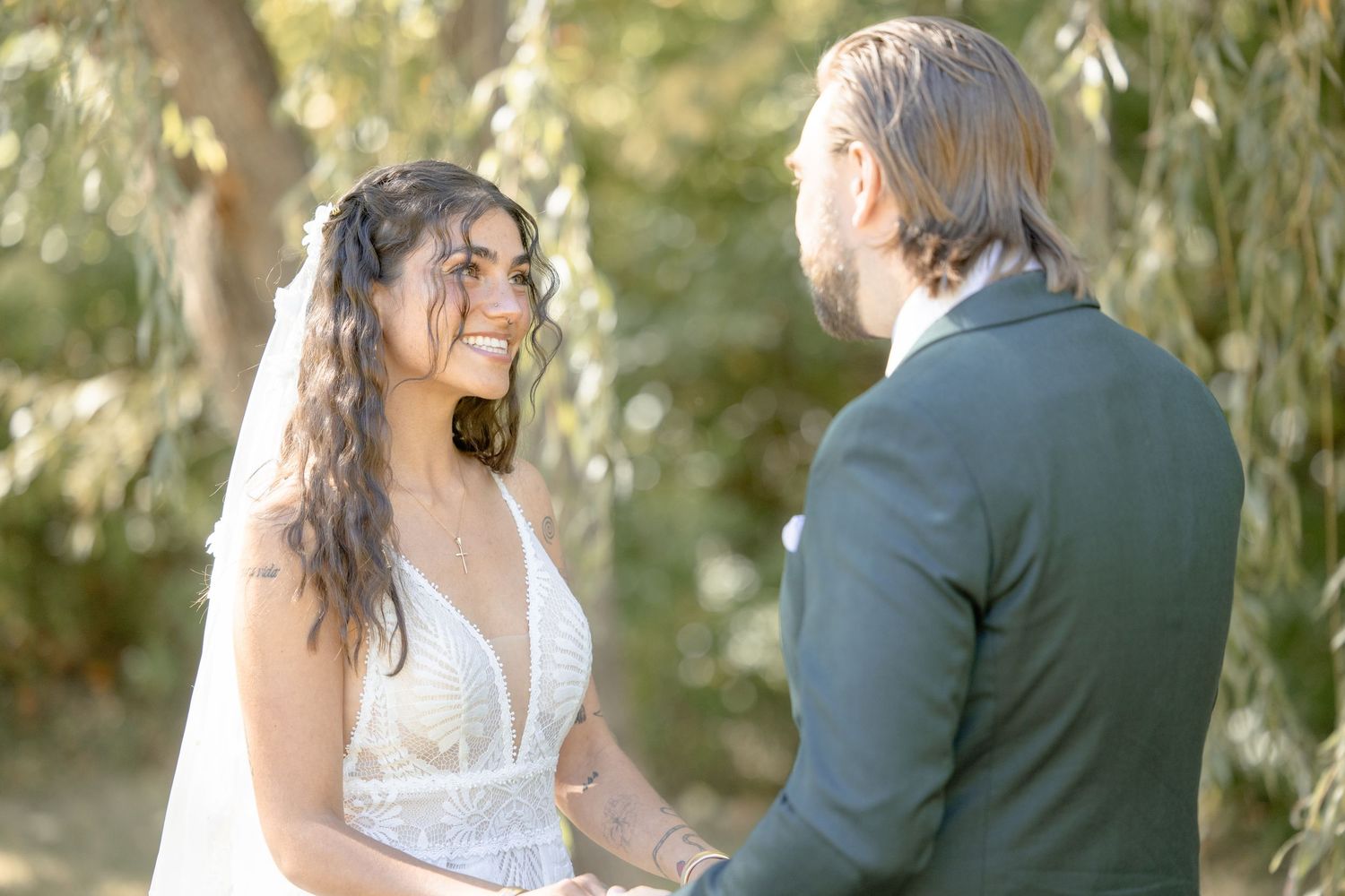 A romantic wedding couple shares tender moments in a garden setting with soft natural lighting and willow trees.