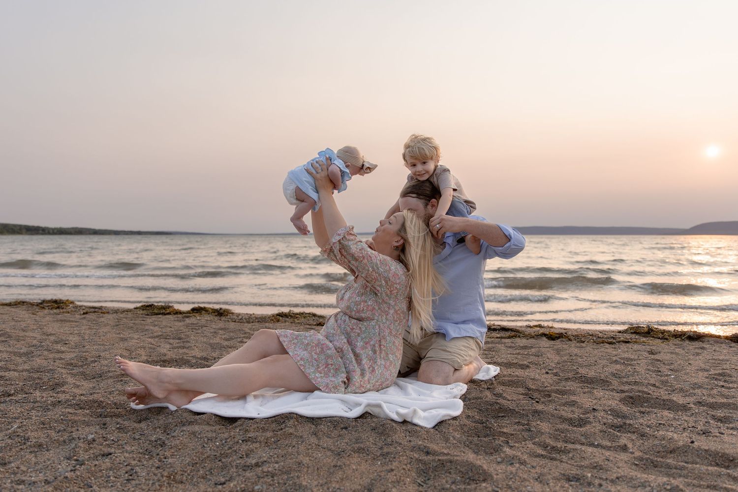 A family plays together on a beach blanket at sunset near the ocean.