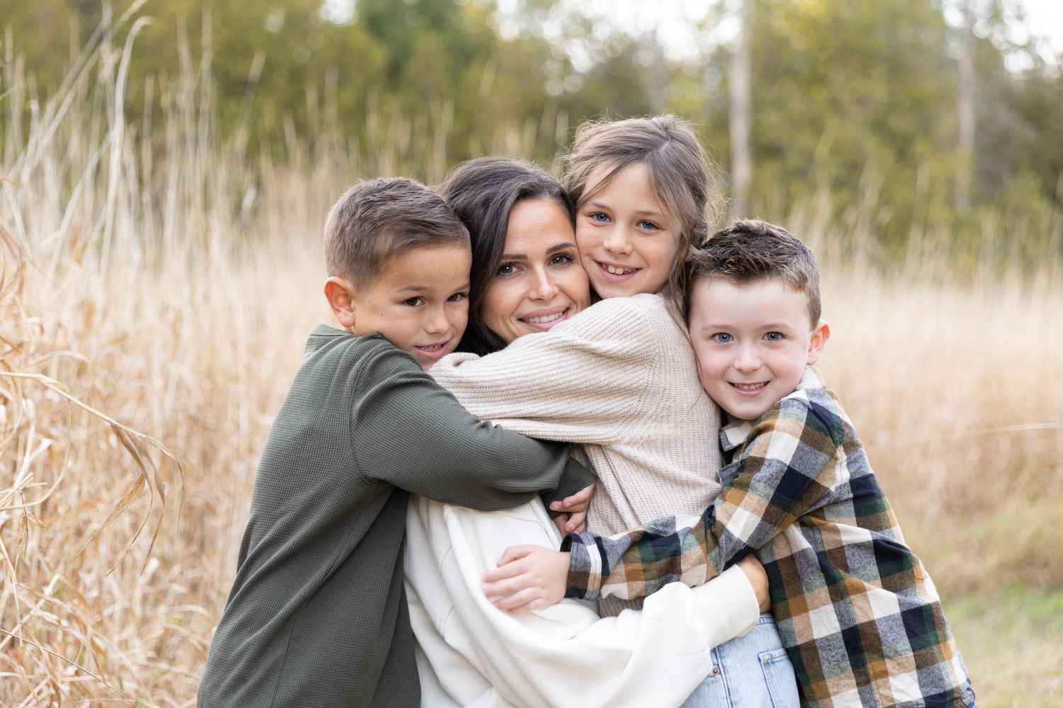 A loving family embraces together while standing in a field of tall dried grass on a sunny autumn day.