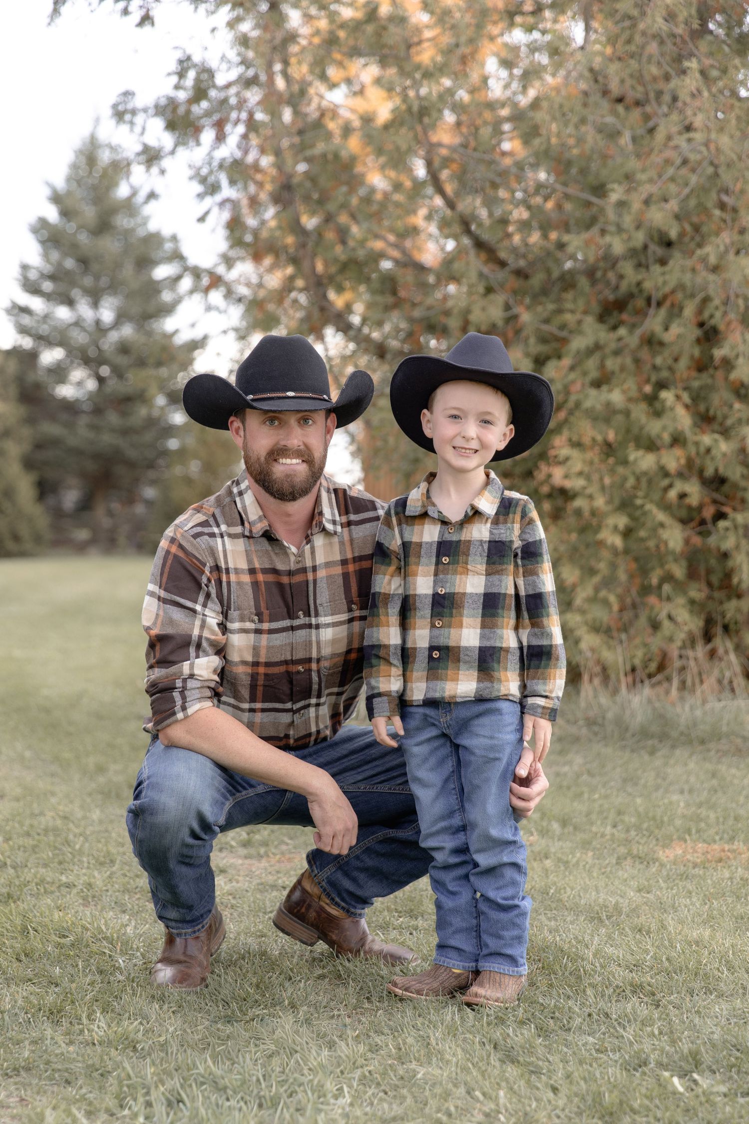 Two figures in matching plaid shirts and cowboy hats kneel together on grassy ground during autumn.