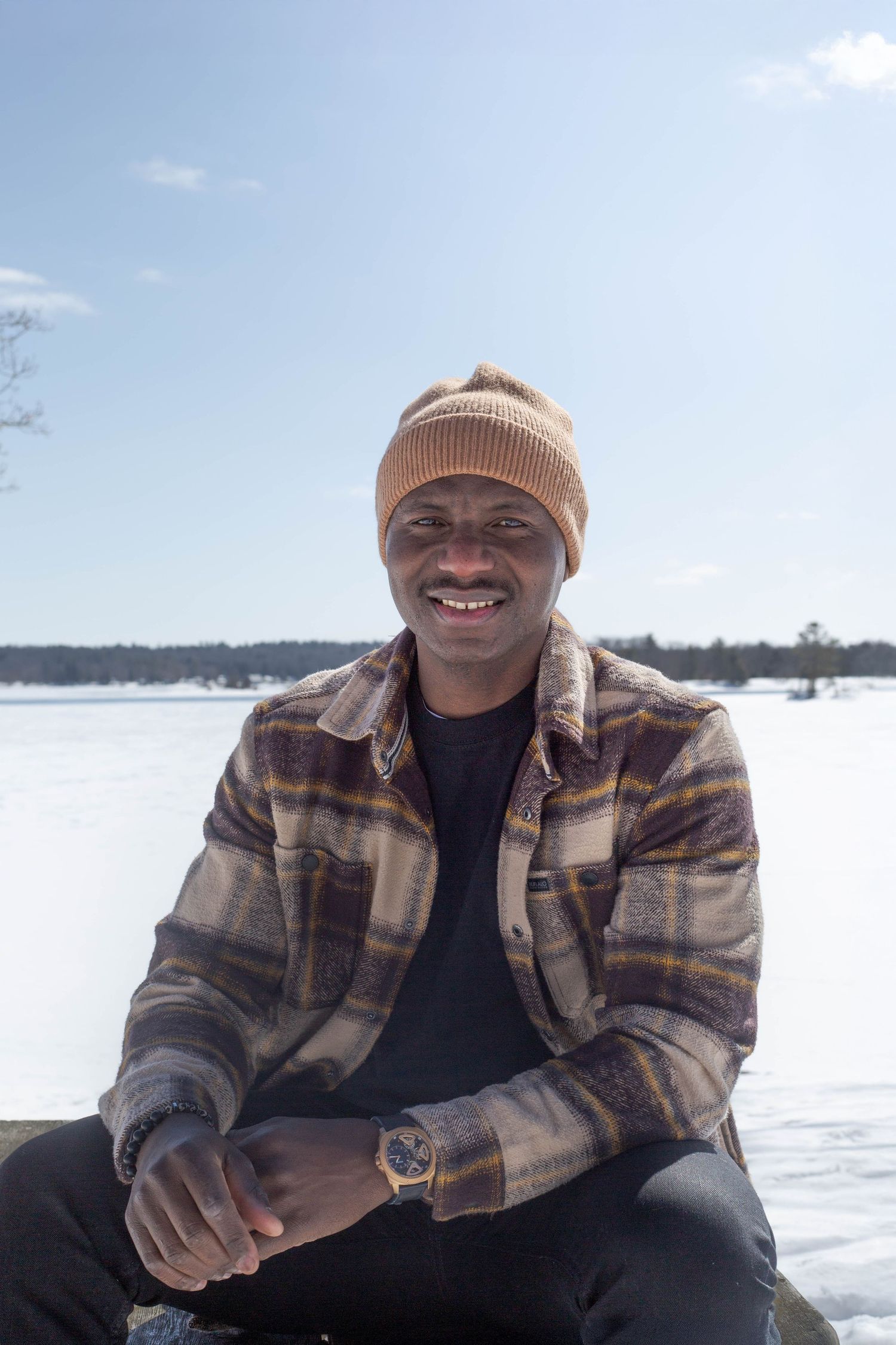 A man wearing a plaid jacket and beanie smiles while sitting on a dock during winter.