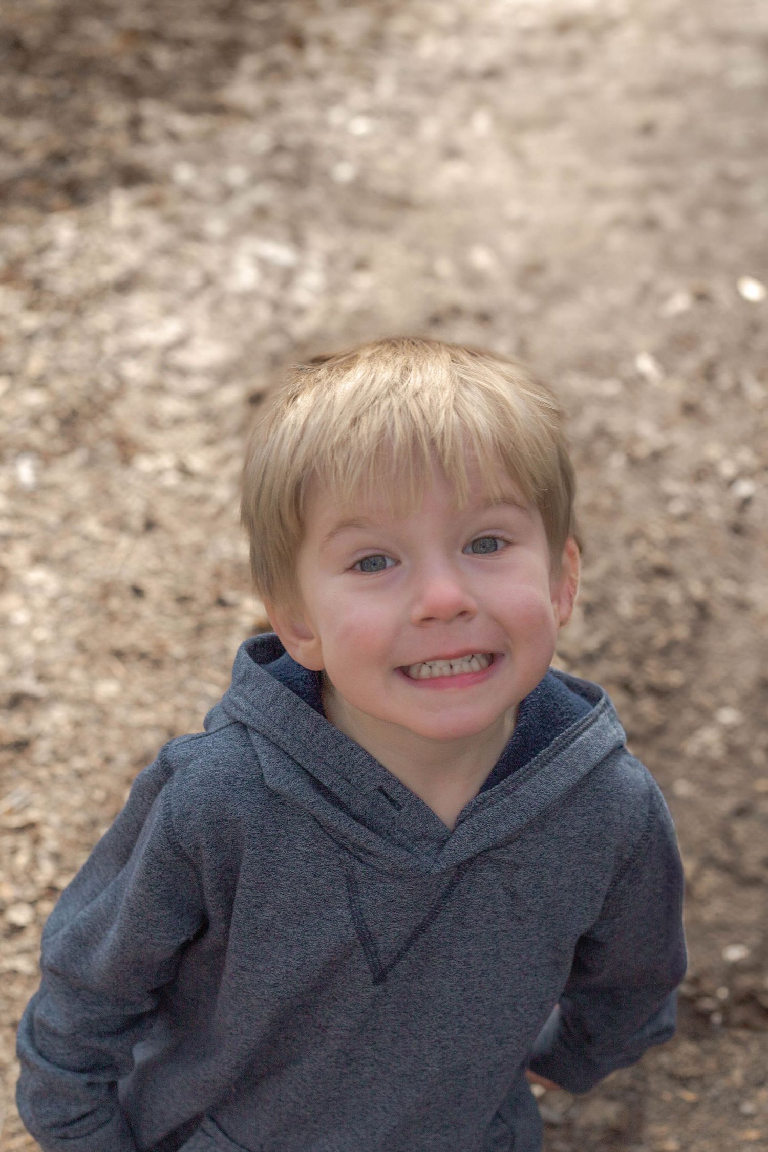 A cheerful child wearing a gray hooded sweatshirt smiles warmly outdoors against a blurred natural background.