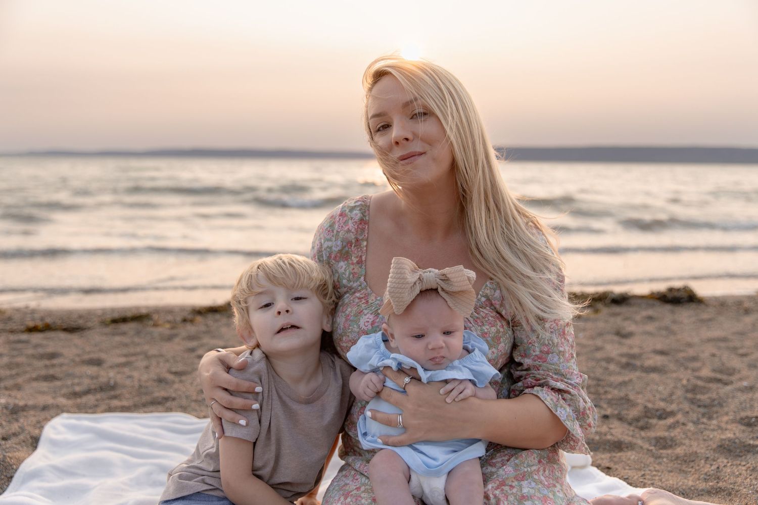A family group sits together on a blanket at the beach during sunset.