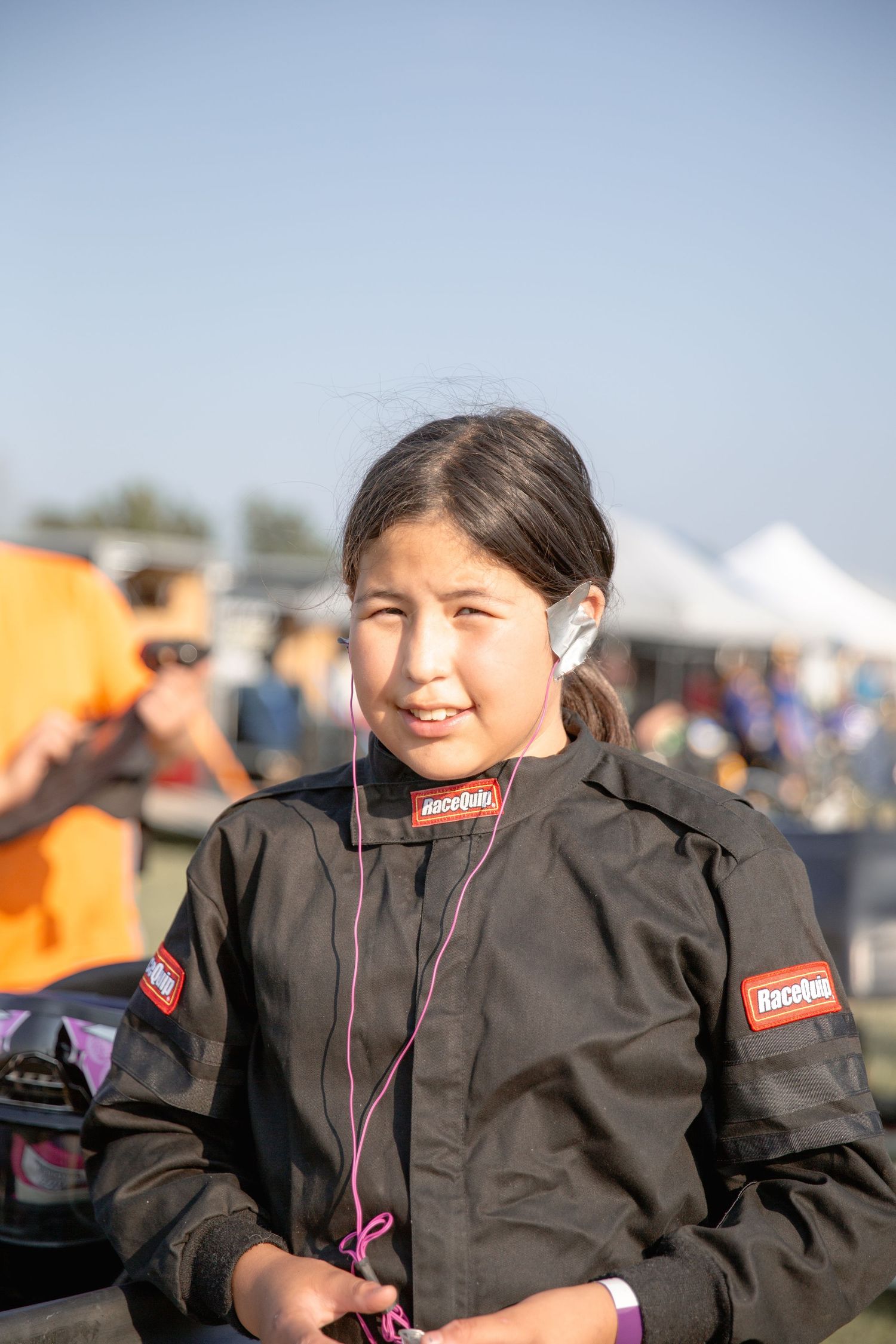 A youth in a black technical jacket with earbuds stands at an outdoor event with people and tents in background.