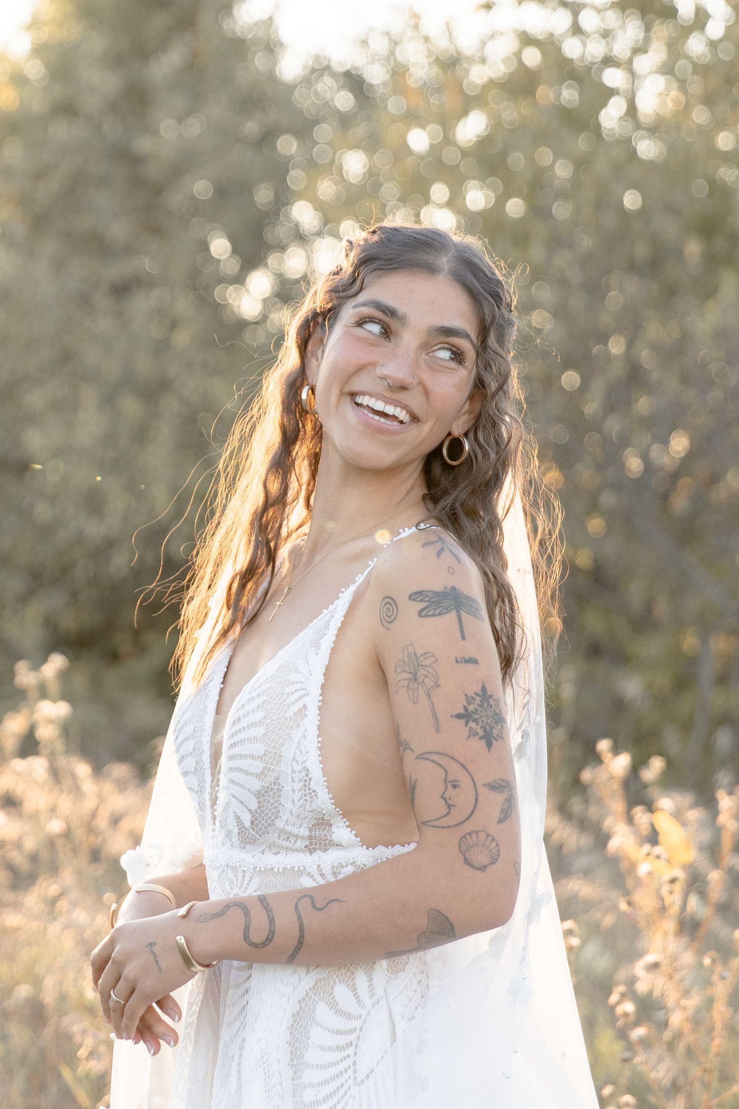 Woman in white lace dress poses outdoors at golden hour with visible tattoos and flowing hair.