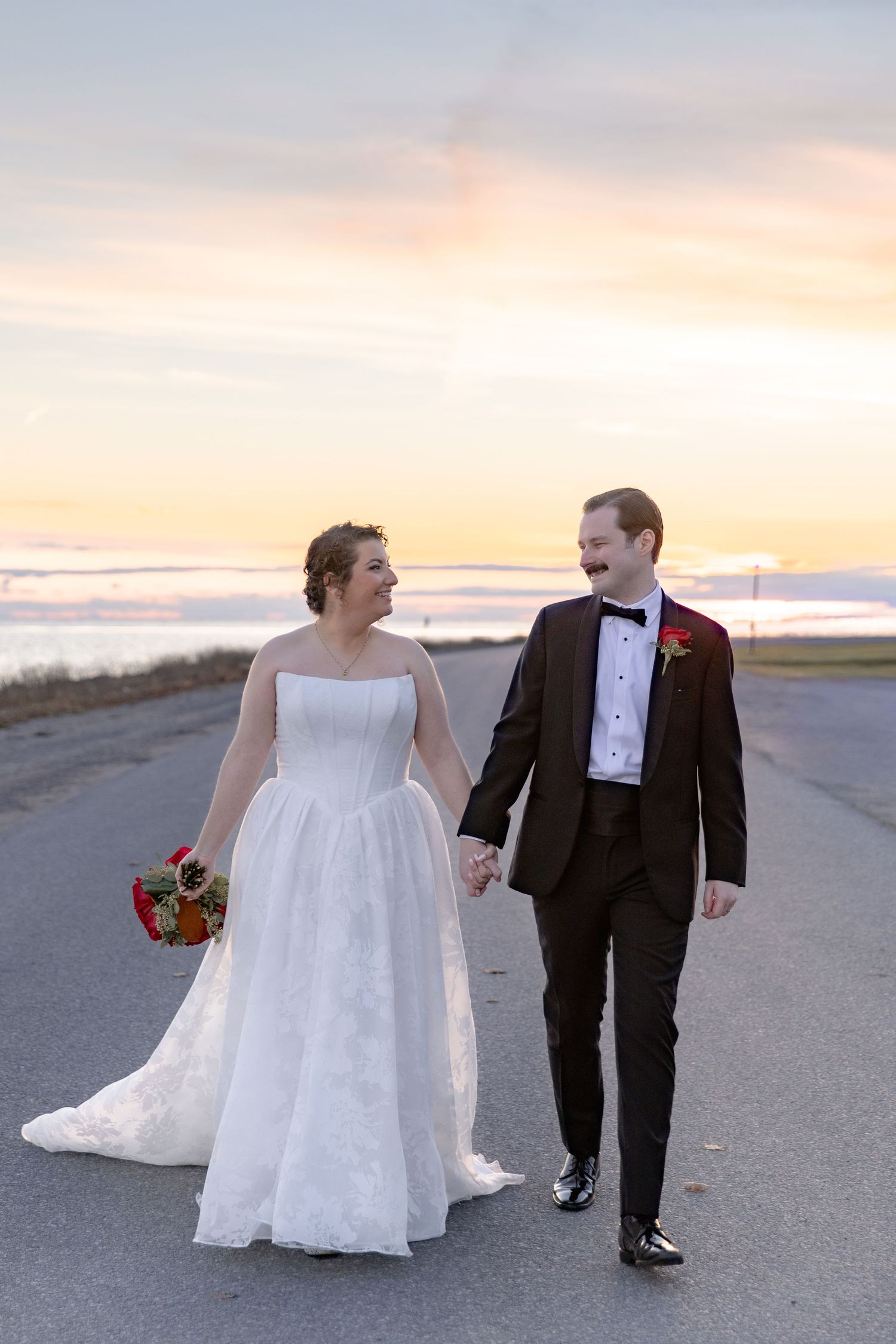 A couple in wedding attire walks along a beach path during a beautiful sunset, creating a romantic silhouette.