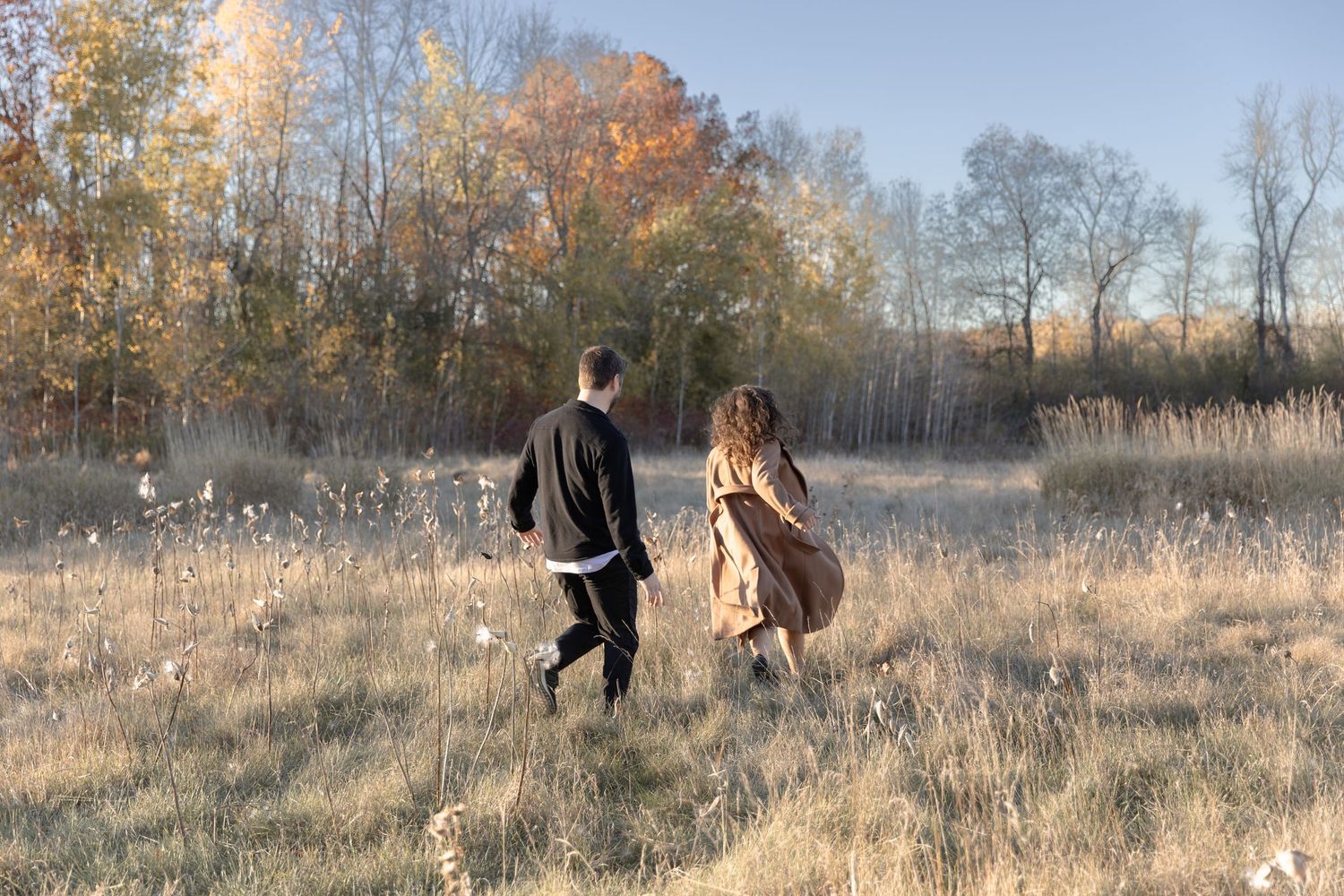 People playing and throwing snow in a sunlit winter field surrounded by bare trees and golden grasses.