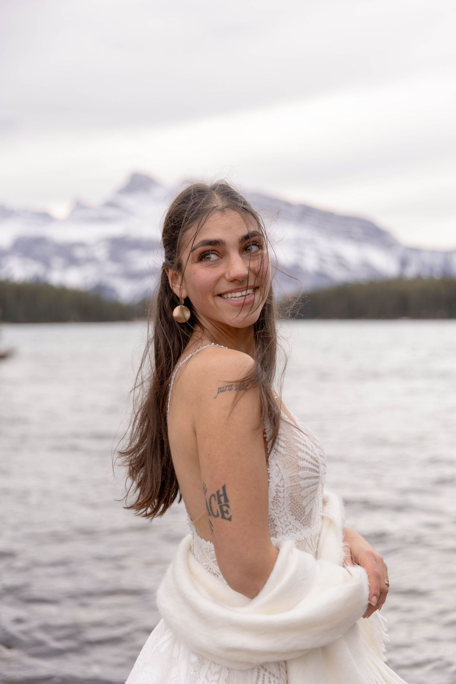 Woman in white wrap poses against snowy mountain landscape with lake in winter setting.
