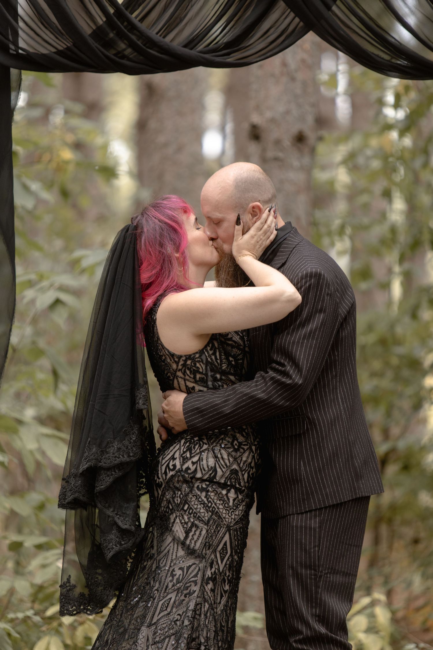 A romantic couple in formal attire share tender moments in a wooded outdoor setting.