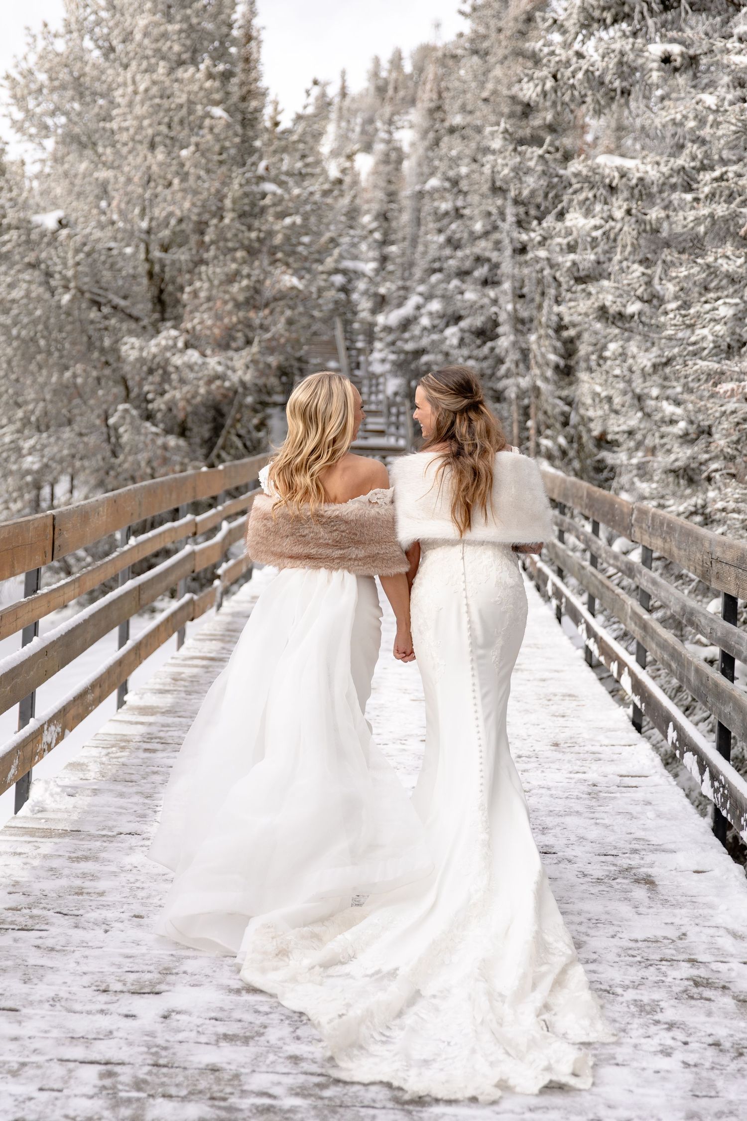 Two women in white dresses walk along a snowy wooden boardwalk surrounded by frost-covered trees in winter.