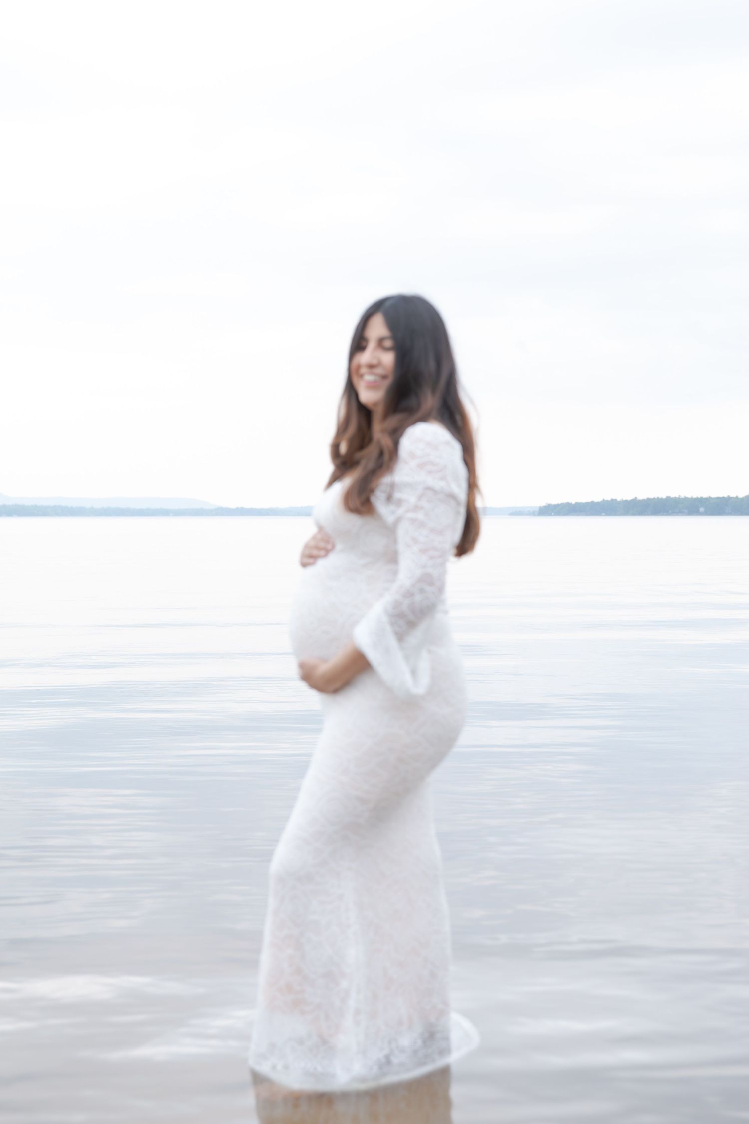 An expectant mother in a white lace dress poses by the water during a maternity photoshoot.