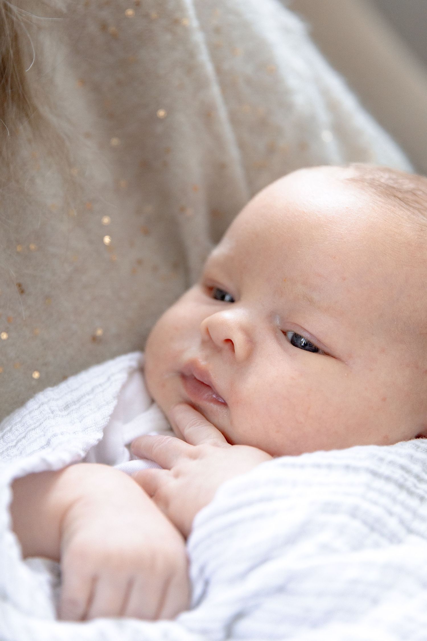 A sweet baby in white clothing rests on soft bedding while gazing upward with curious eyes.