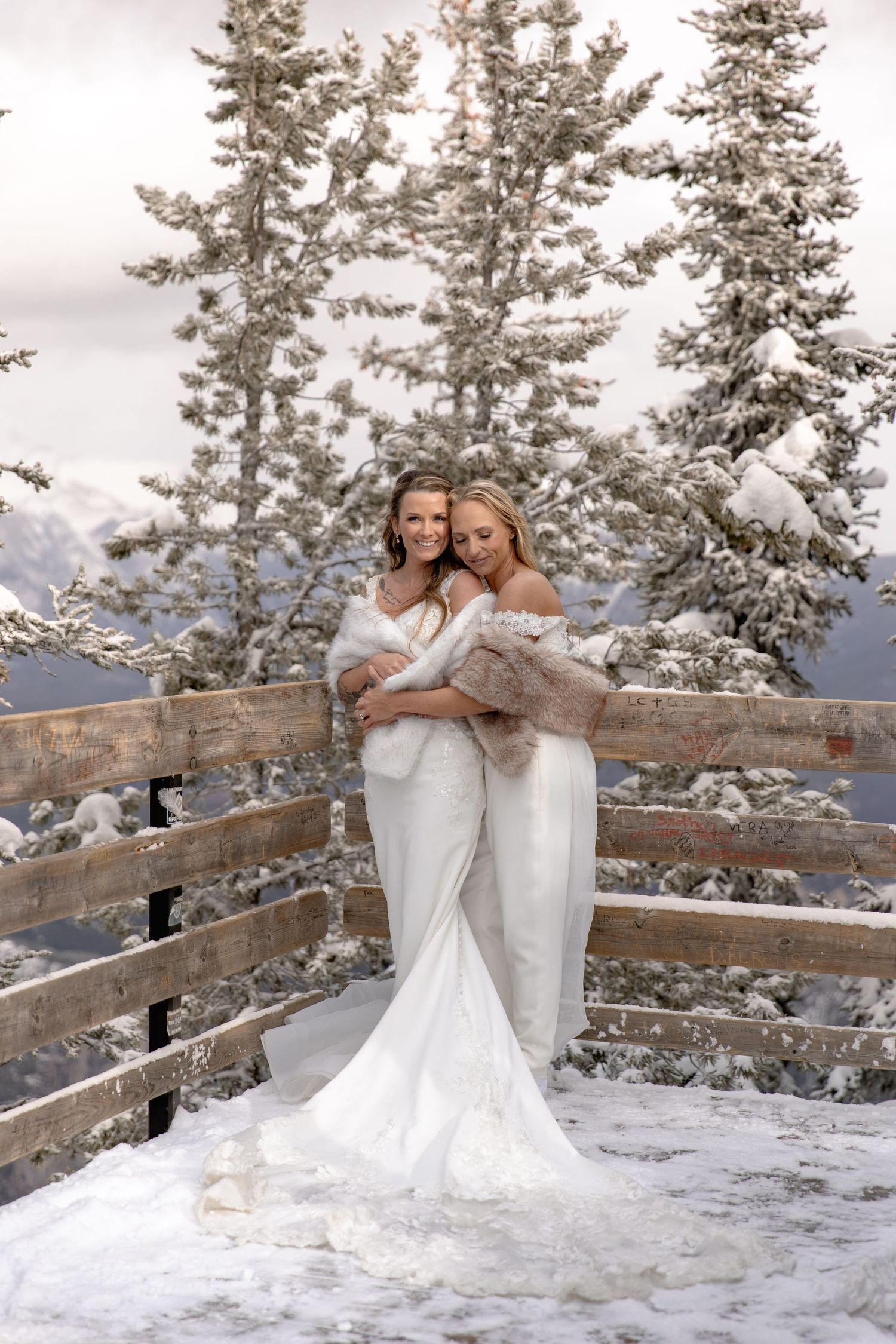 Two brides in white wedding dresses pose together in a snowy winter mountain setting with evergreen trees and wooden fence.