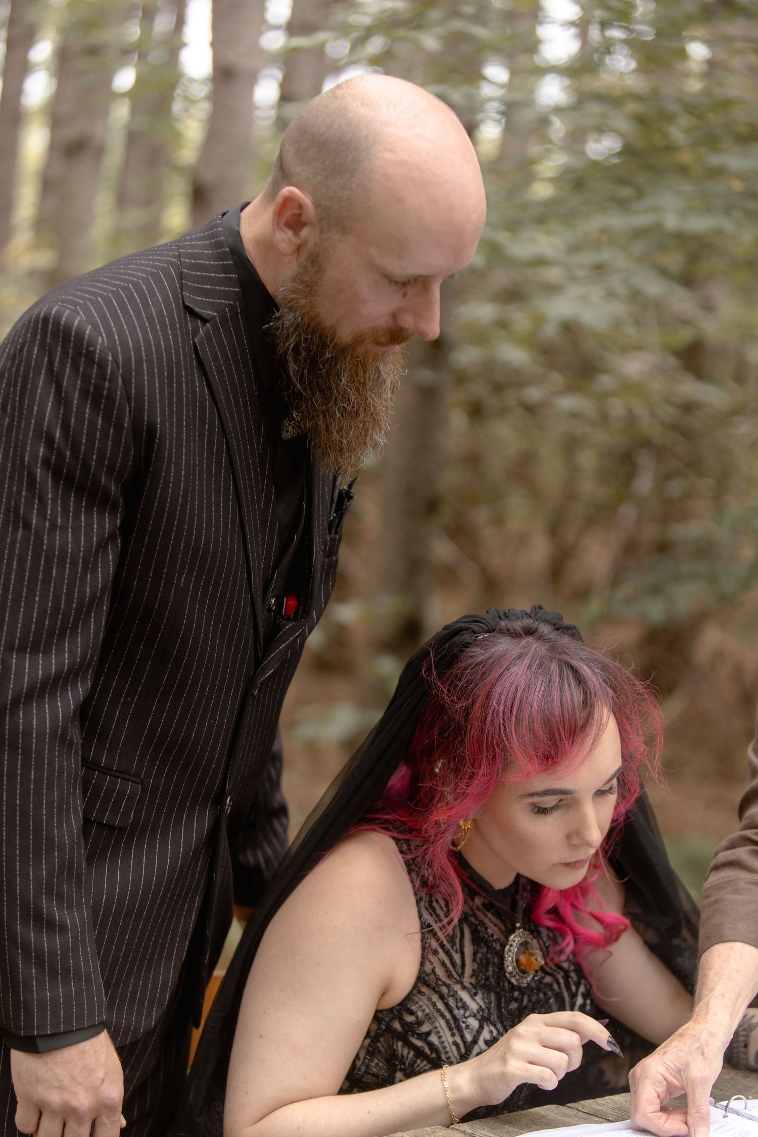 A couple in formal attire examines something together outdoors in a wooded setting.
