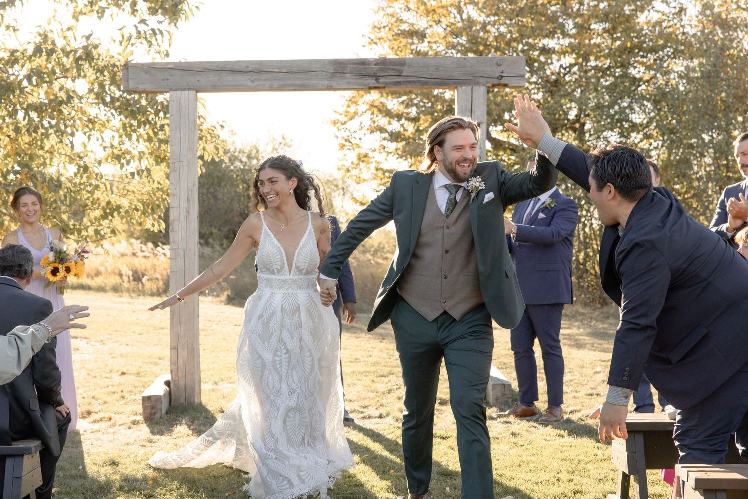 Wedding party celebrates outdoors during golden hour with joyful dancing and laughter in a natural setting.