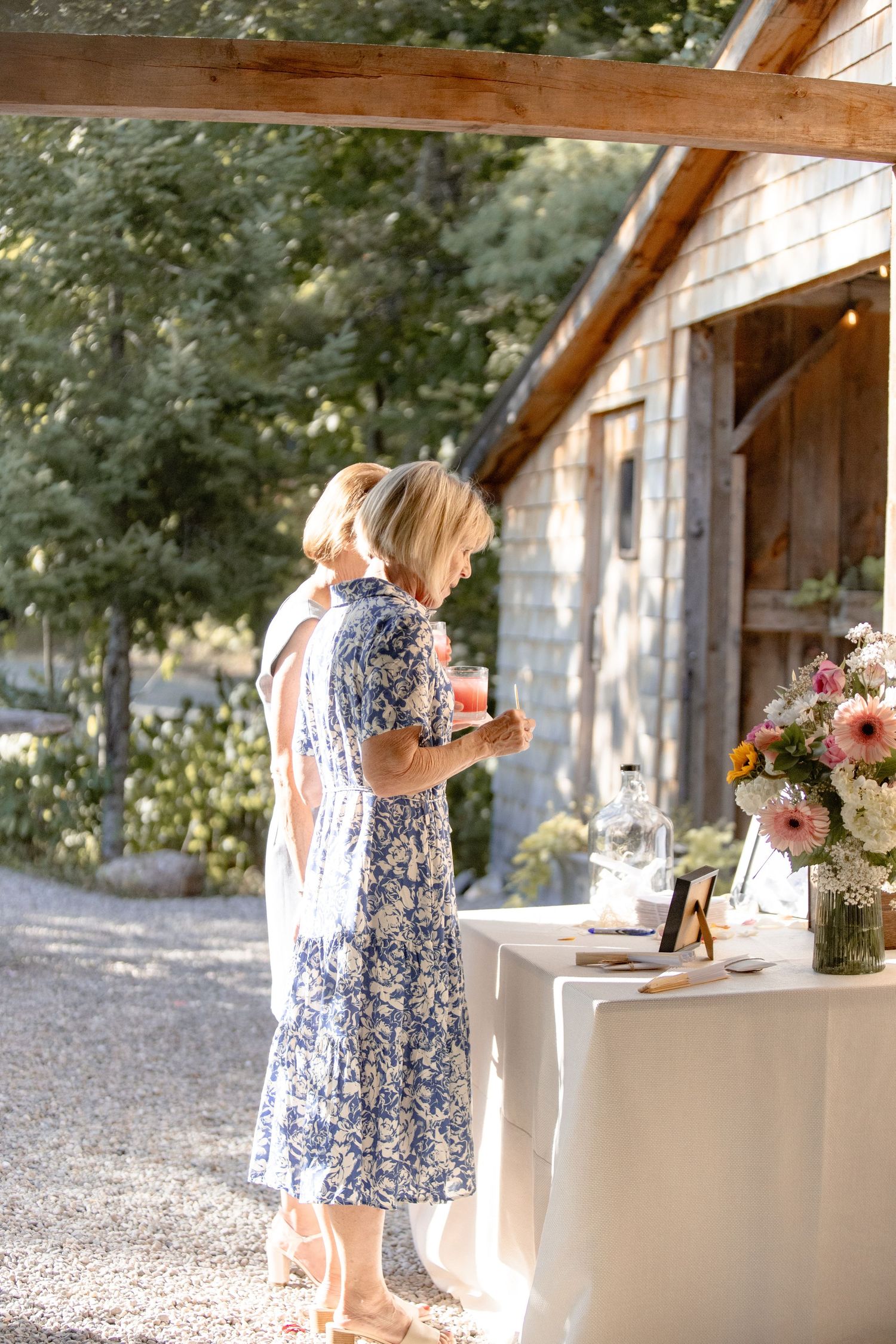 A woman in a blue floral dress arranges flowers at a white table under a rustic wooden covered patio.