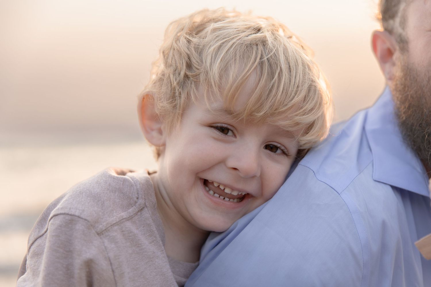 A child with blonde hair leans against an adult wearing a blue shirt on the beach at sunset.