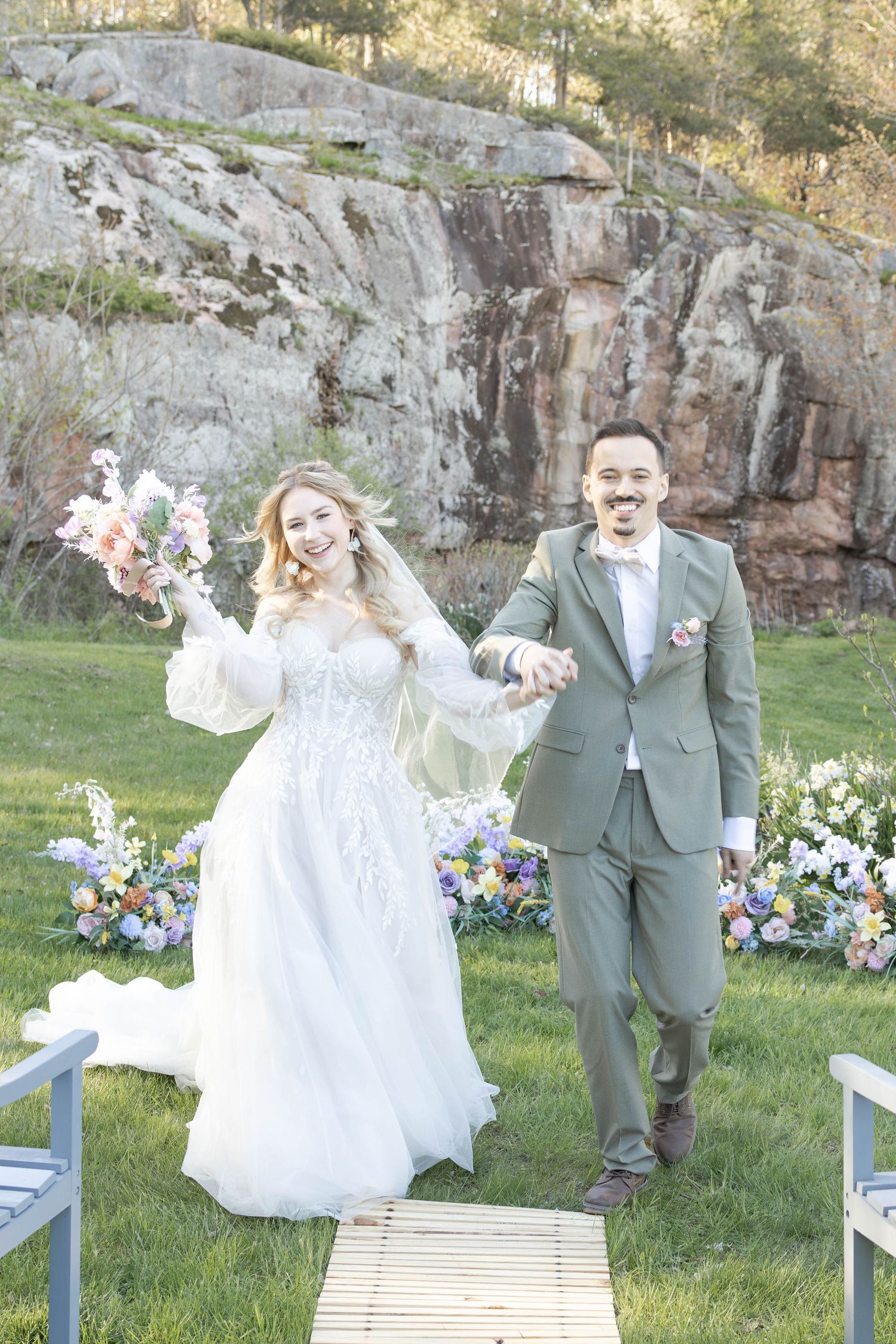 A newlywed couple celebrates joyfully on their wedding day with a rocky cliff backdrop and floral arrangements.