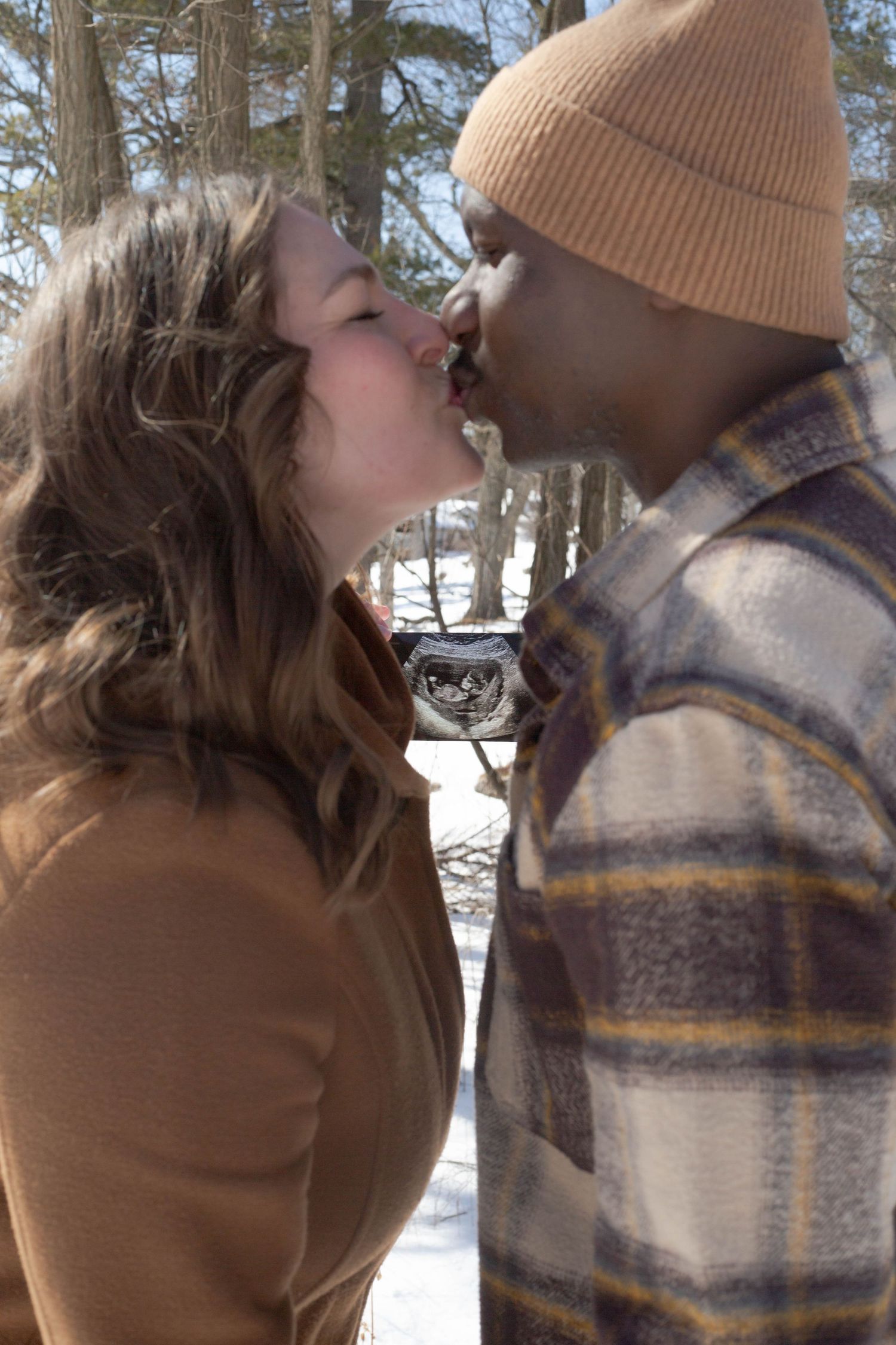 A romantic winter kiss between a couple wearing warm coats in a snowy forest setting and holding an ultrasound image.