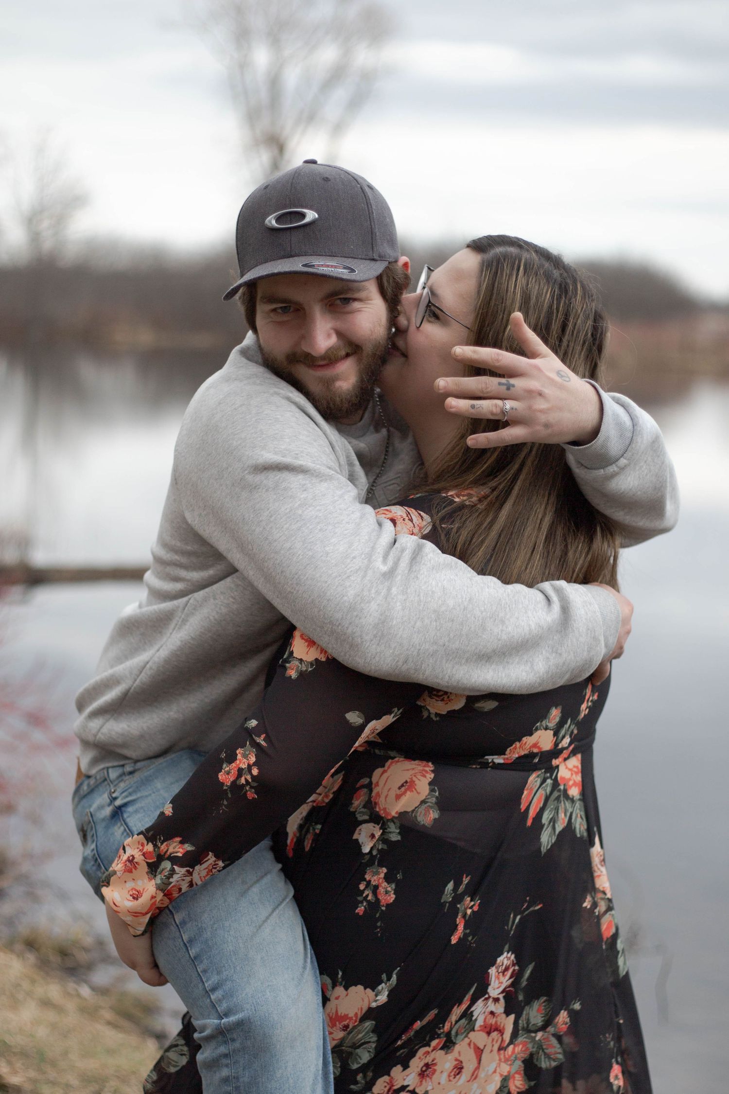 A couple shares a tender playful moment by a lake, with the person in a baseball cap hugging the other wearing a floral dress.