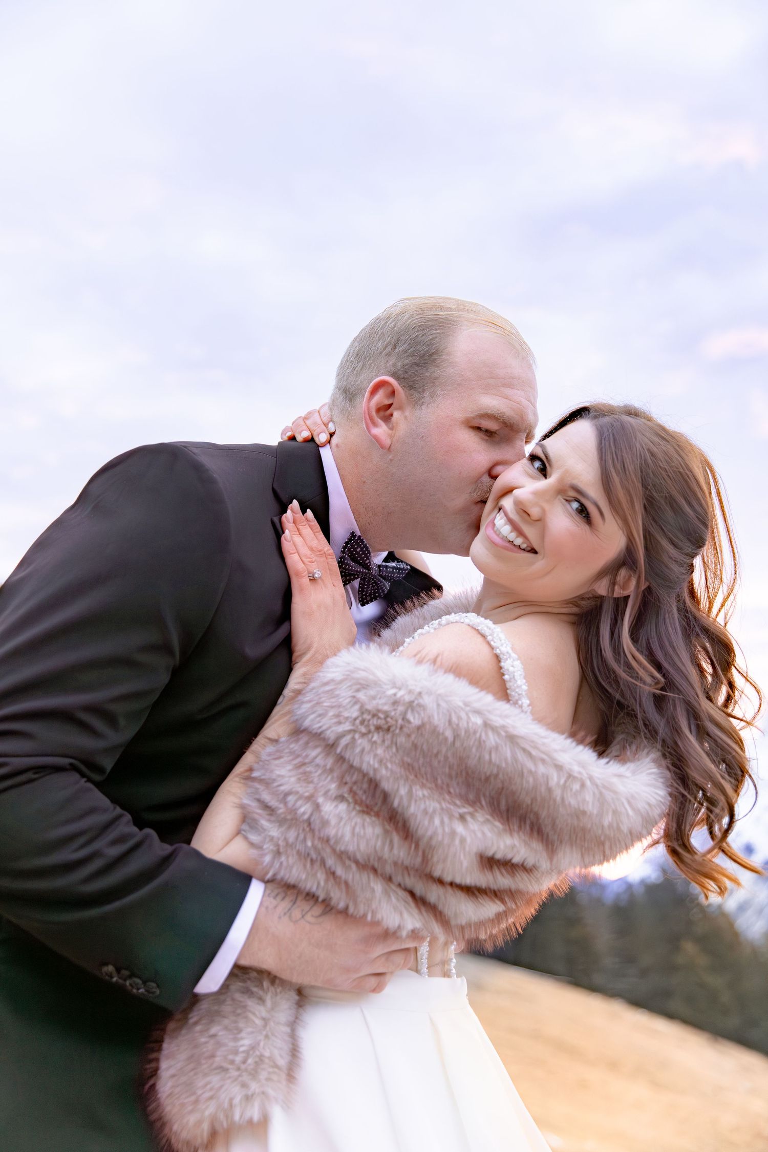 A romantic couple embraces and kisses during a sunset photoshoot, with the bride wearing a fluffy pink jacket.