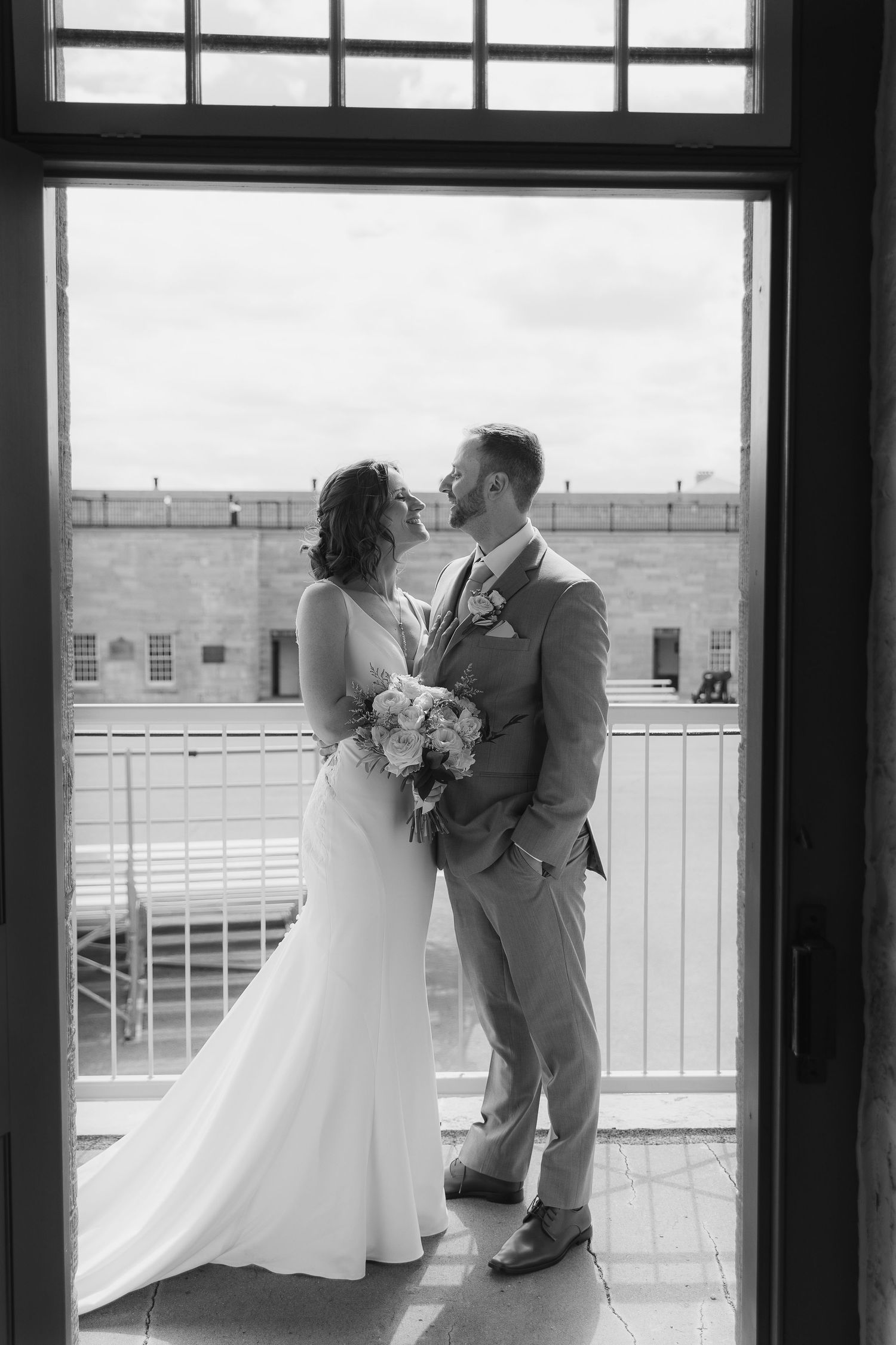 Black and white photo of bride and groom sharing a romantic moment in a doorway, bride holding white bouquet.
