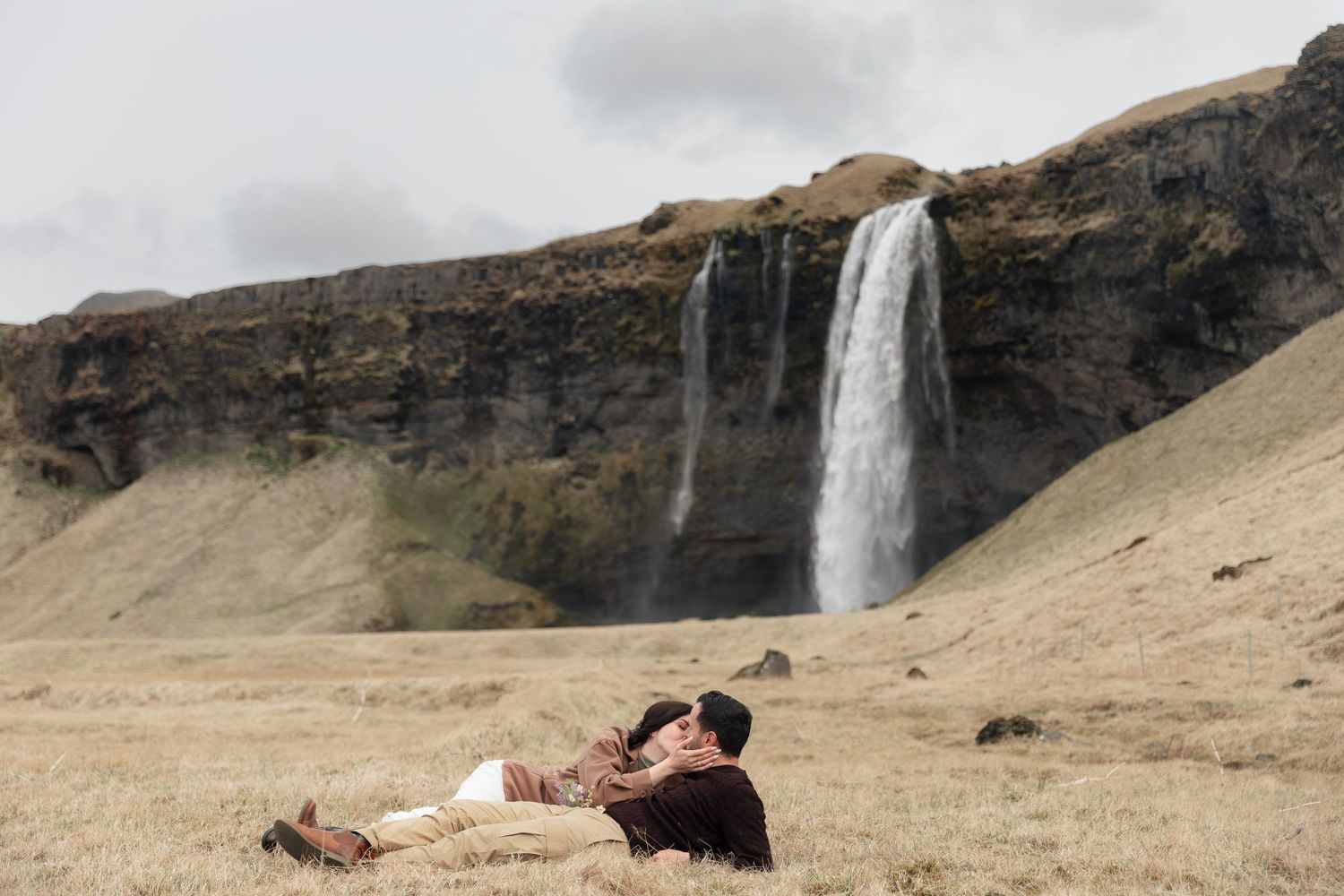 Couple lying in golden grass fields with Seljalandsfoss waterfall cascading down dramatic cliffs in Iceland.