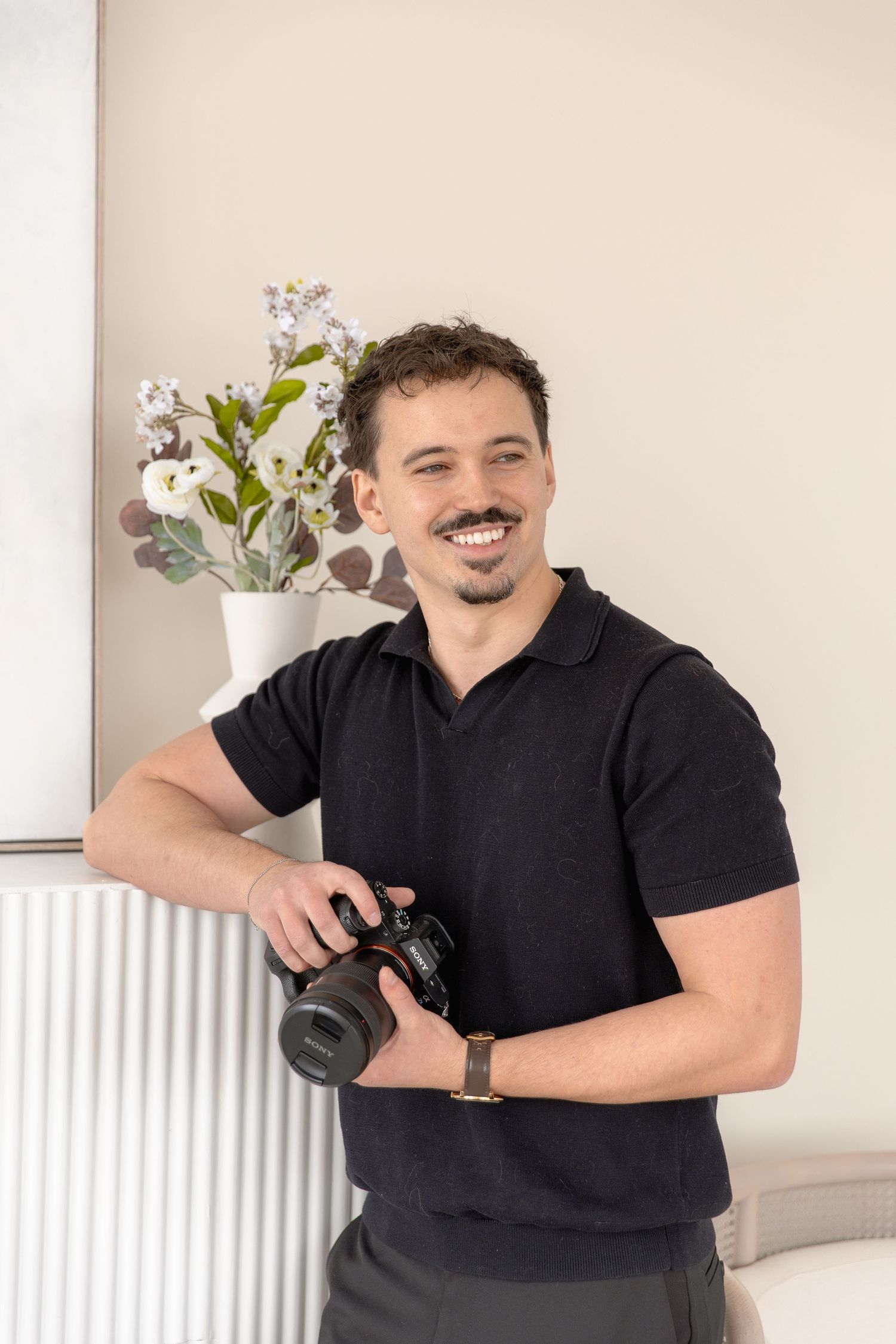 Photographer in black polo shirt smiles while holding camera in bright interior setting with white flowers.